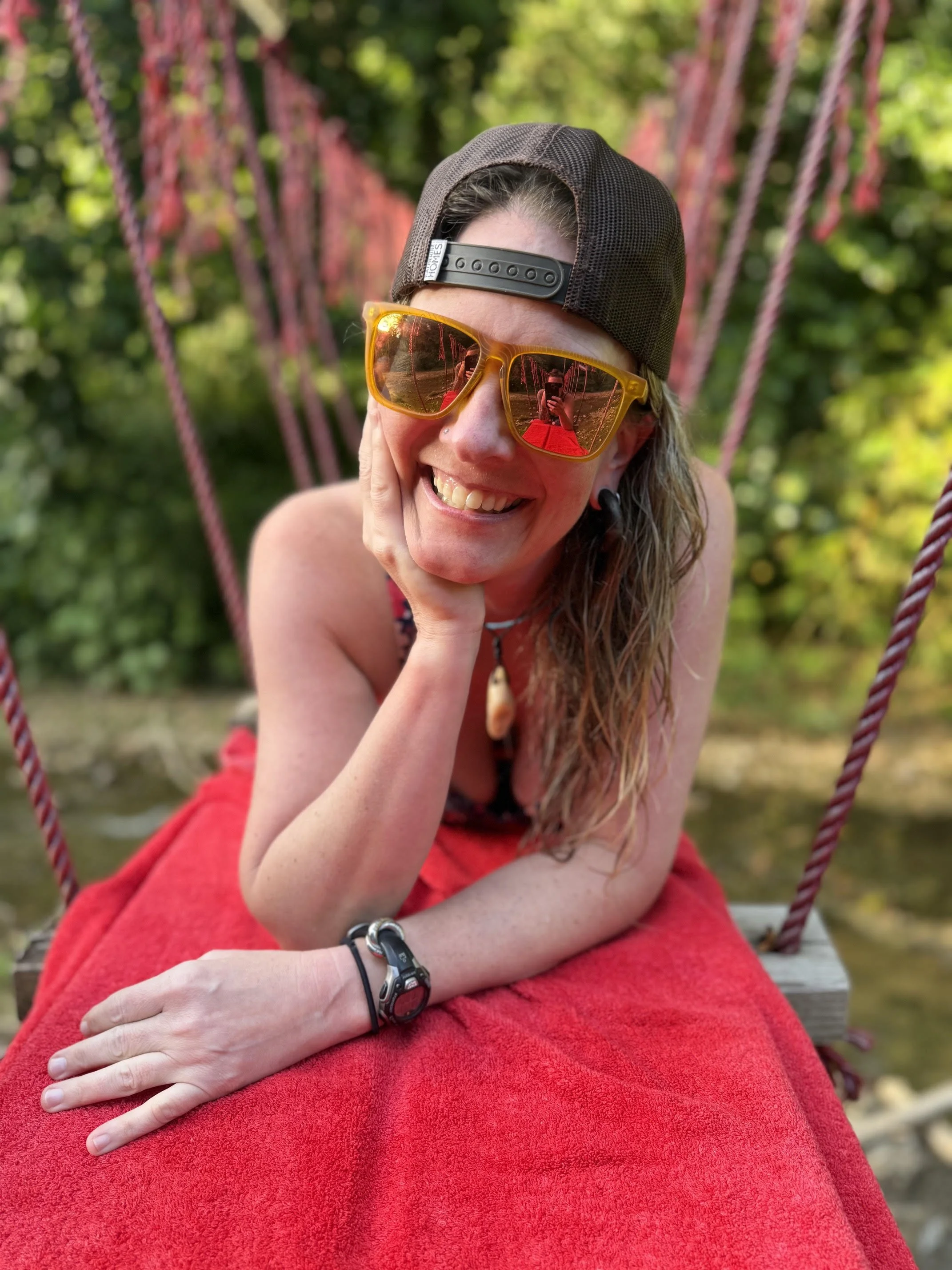 A woman with long curly hair wearing an orange-tinted sunglasses, a black cap backwards, and a red dress, sitting on a swing outdoors surrounded by green foliage.