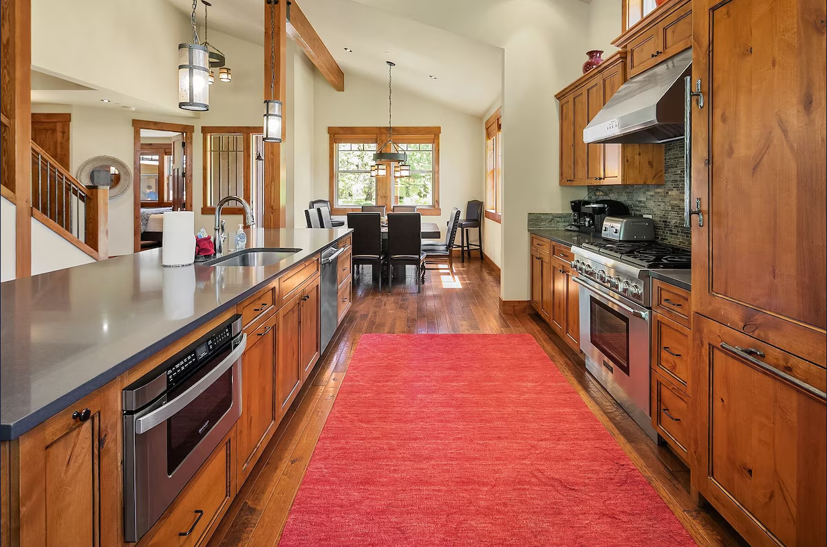 Kitchen with wooden cabinets, stainless steel appliances, and a red rug on hardwood floor, with a dining area and windows in the background.