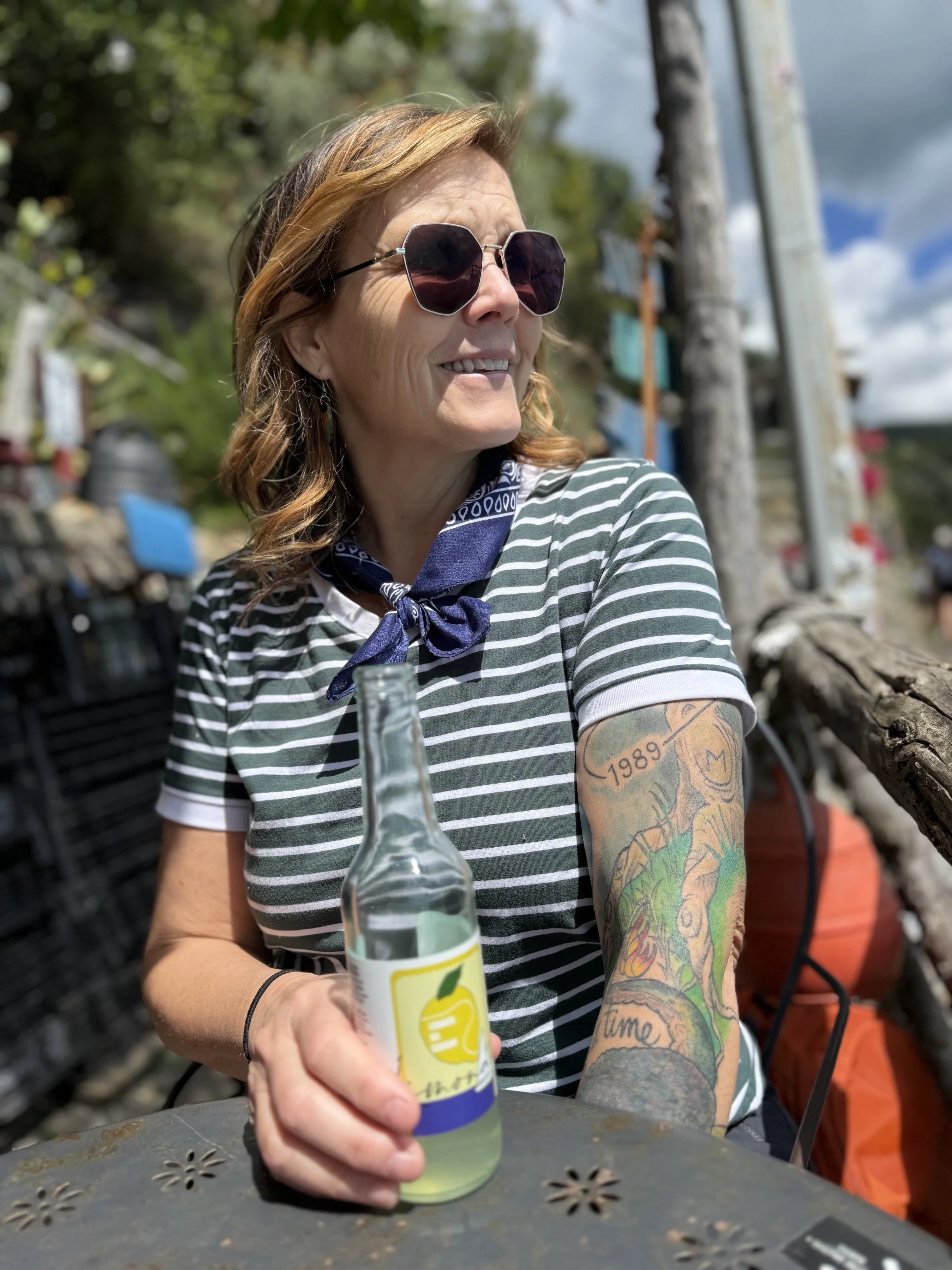 A woman with orange hair, wearing sunglasses, a striped shirt, and a blue bandana around her neck, sitting outdoors at a table, holding a clear bottle with a lemon logo, smiling and looking away from the camera.