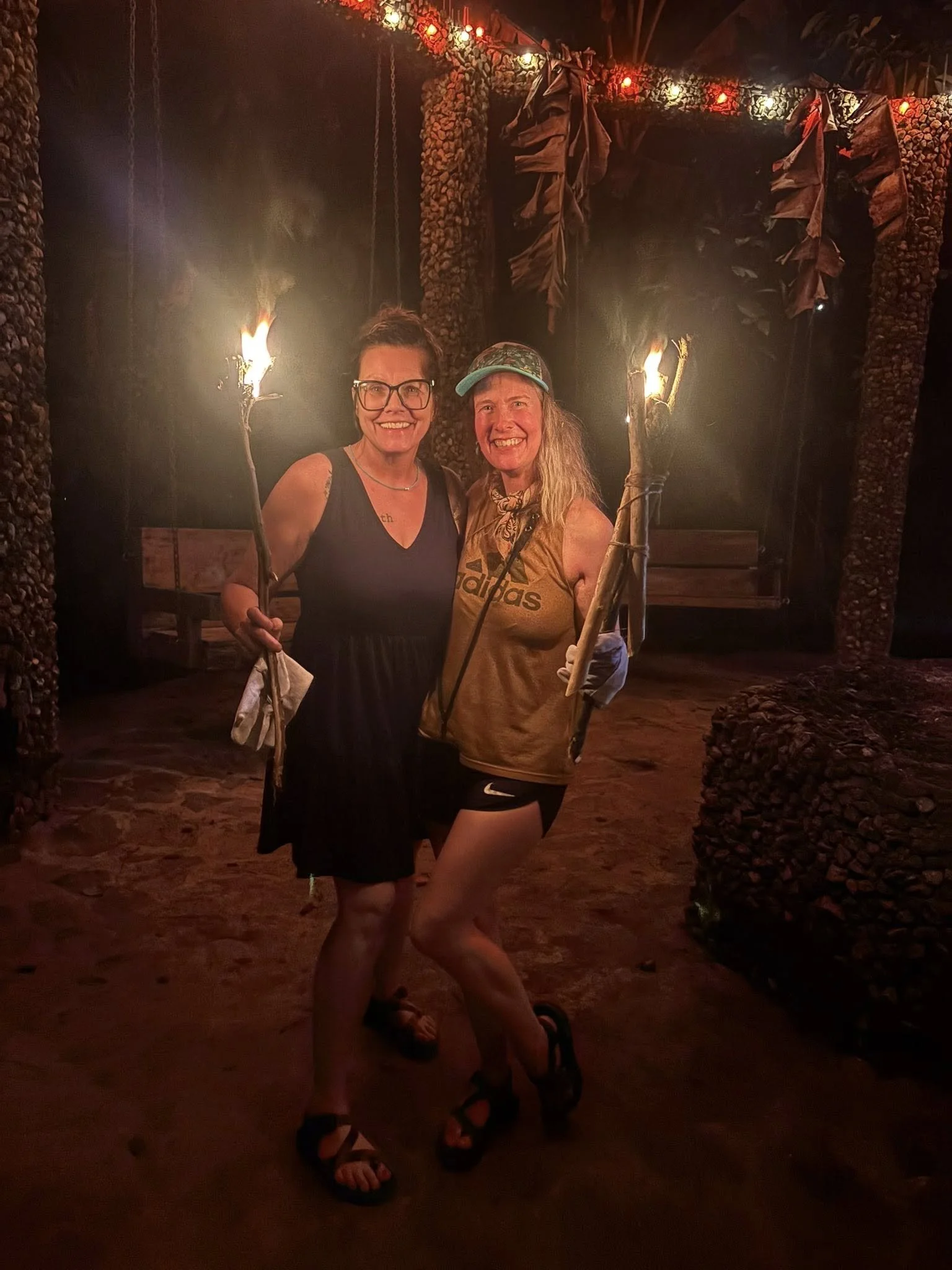 Two women smiling and holding tiki torches at night, standing inside a decorative archway with stone and leaf elements, illuminated by string lights.