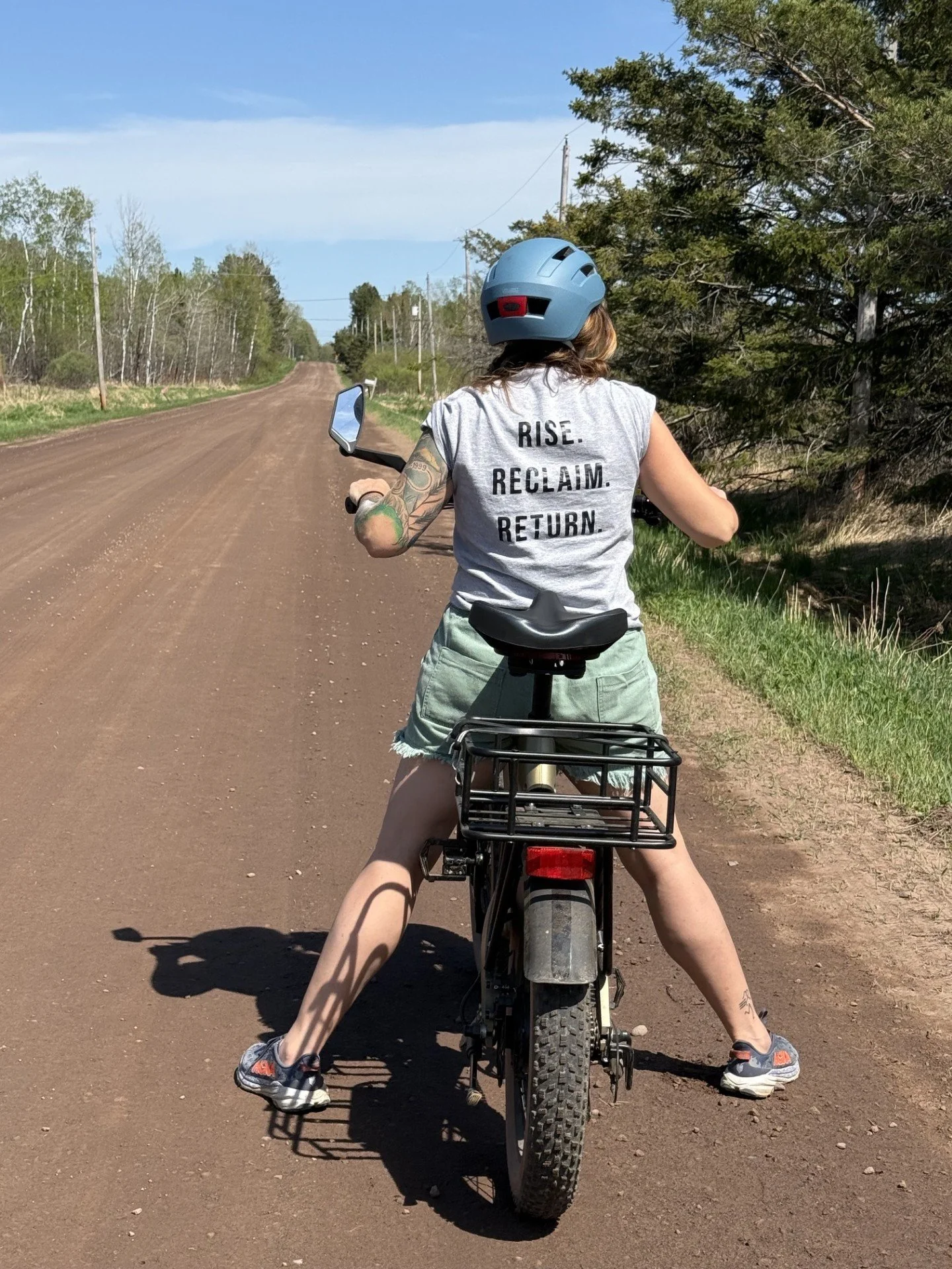 Person riding a bicycle on a rural dirt road with trees and power lines in the background, wearing a light blue helmet and a sleeveless shirt with the words "RISE. RECLAIM. RETURN." on the back, and shorts.