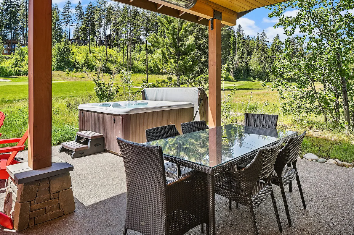 Patio with a glass-top dining table and six black wicker chairs, surrounded by greenery including trees and a field, with a hot tub to the back and a covered roof overhead.
