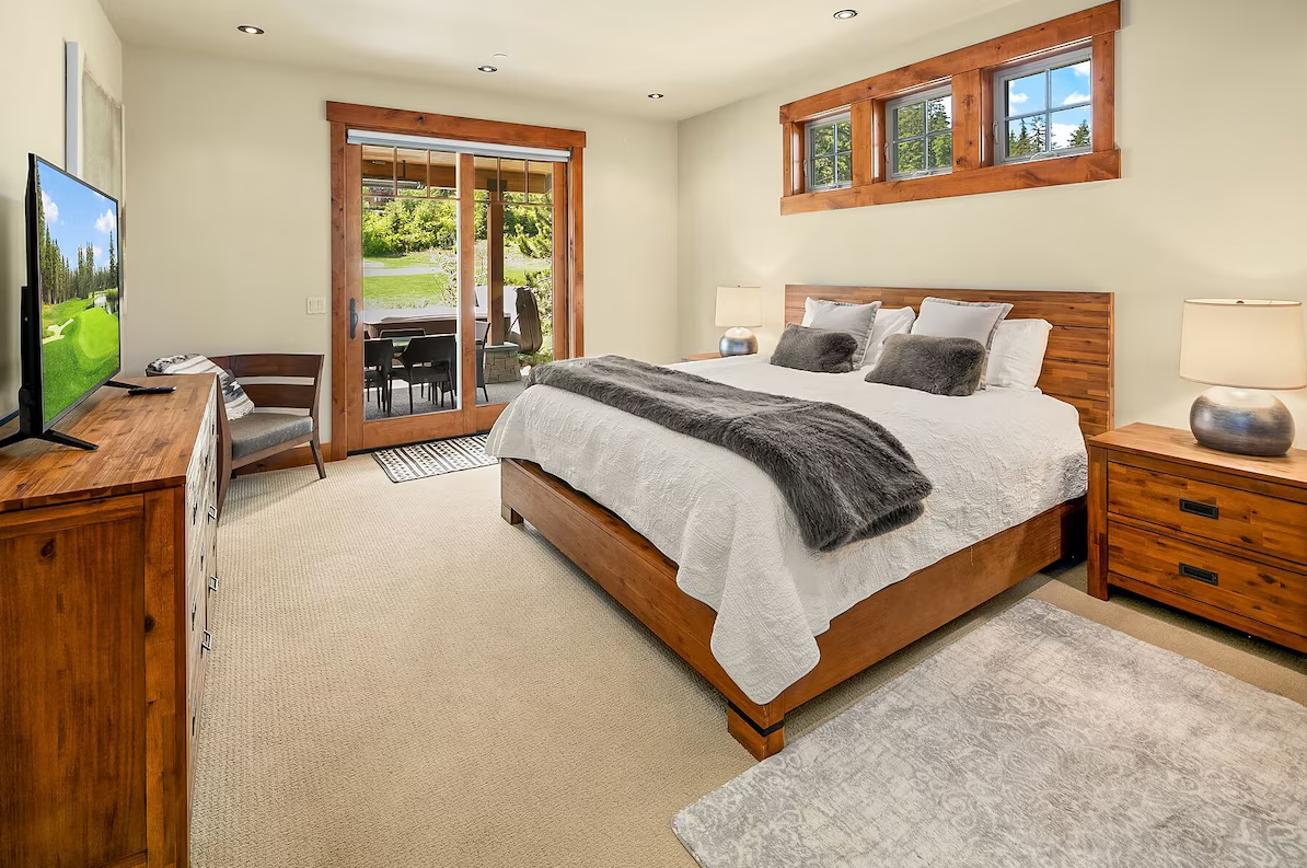 A bedroom with a large wooden bed, white bedding, and gray accent pillows. There are two wooden nightstands with lamps, a wooden dresser with a television, and a sliding glass door leading to an outdoor patio. Windows above the bed bring in natural l