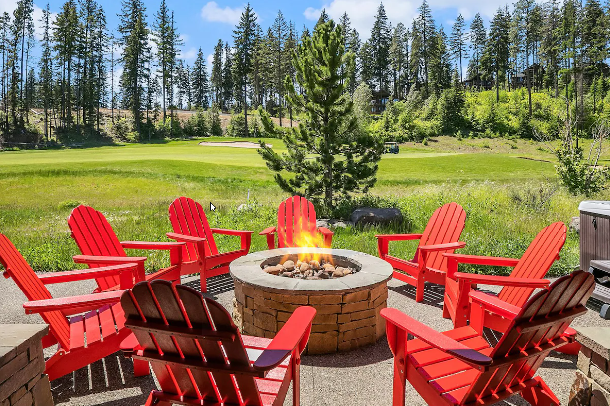 Six red Adirondack chairs arranged around a stone fire pit on a patio with a grass field, trees, and a blue sky in the background.