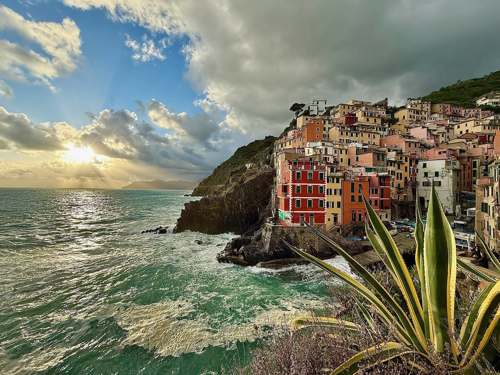 Colorful hillside houses overlooking the ocean with a partly cloudy sky and the sun setting on the horizon.