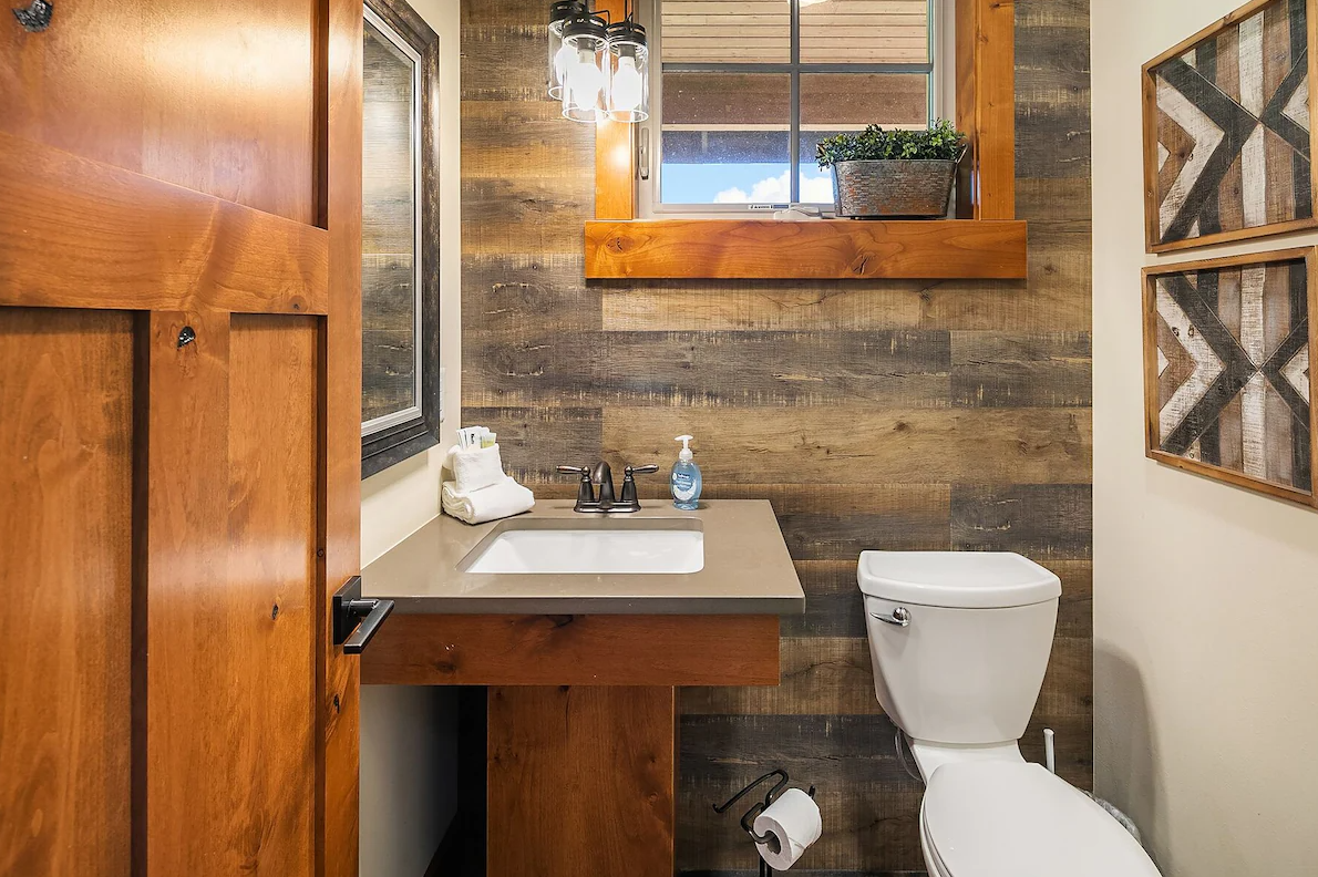 A rustic bathroom featuring a white toilet, a small sink with a beige countertop, a wooden wall panel behind the sink, a window with a flower box, and decorative wall art.