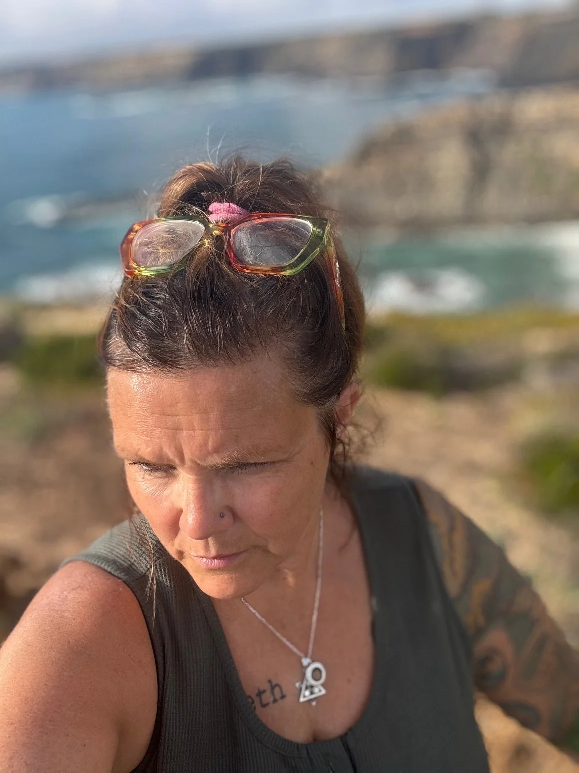 A woman with dark hair, sunglasses on her head, and a nose piercing, looking downward with her eyes closed, standing outdoors near the water with cliffs in the background.