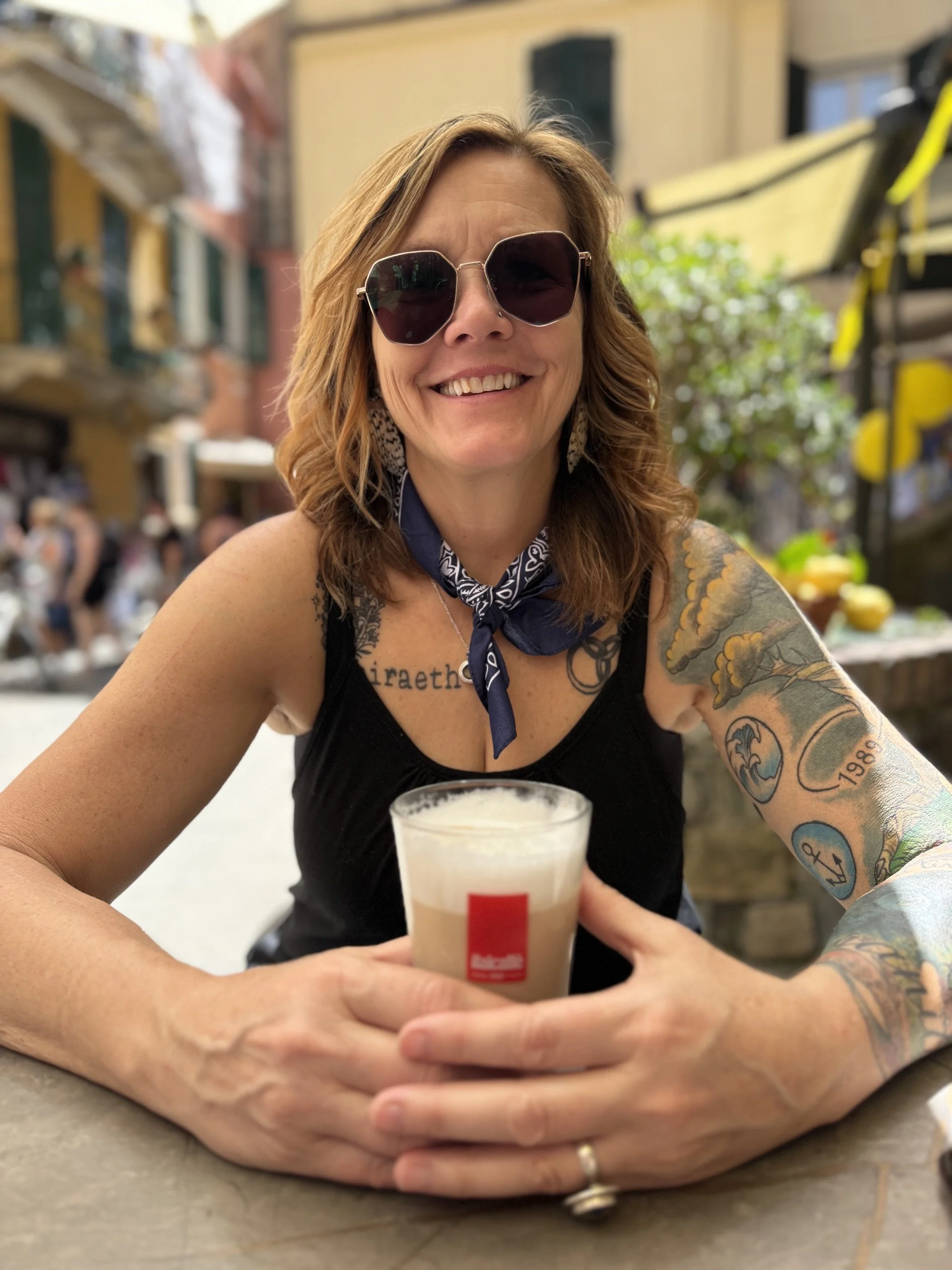 Smiling woman with tattoos, sunglasses, and a tie scarf holding a glass of beer at a lively outdoor event.
