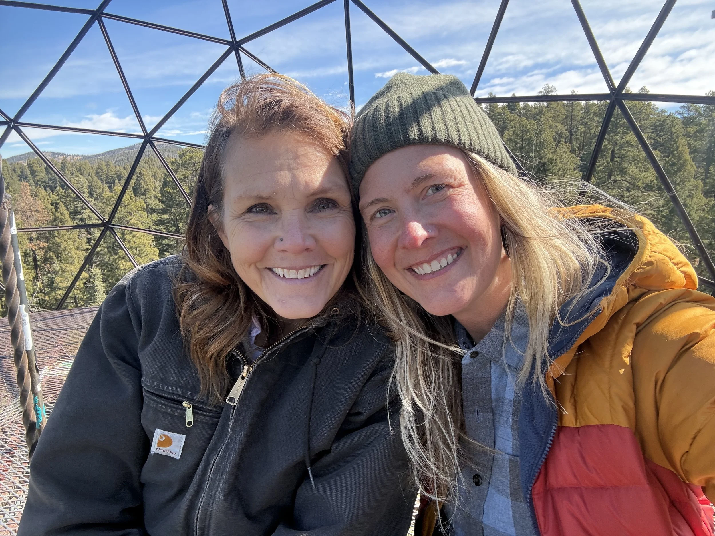 Two women smiling and taking a selfie outdoors with a metal geodesic dome structure above them, surrounded by green trees and mountains under a partly cloudy sky.