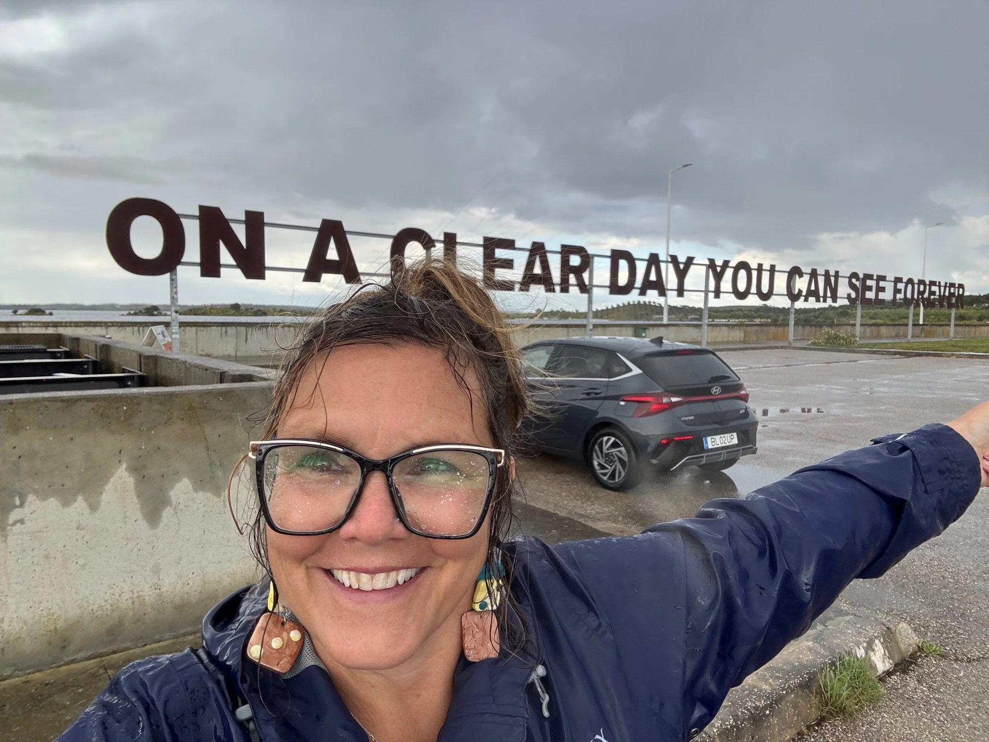 A smiling woman with glasses and earrings taking a selfie outdoors on a rainy day. She is wearing a blue jacket. In the background, there is a large sign that reads 'ON A EAR DAY YOU CAN SEE FOR EVER,' a parking lot with a car, and a cloudy sky.