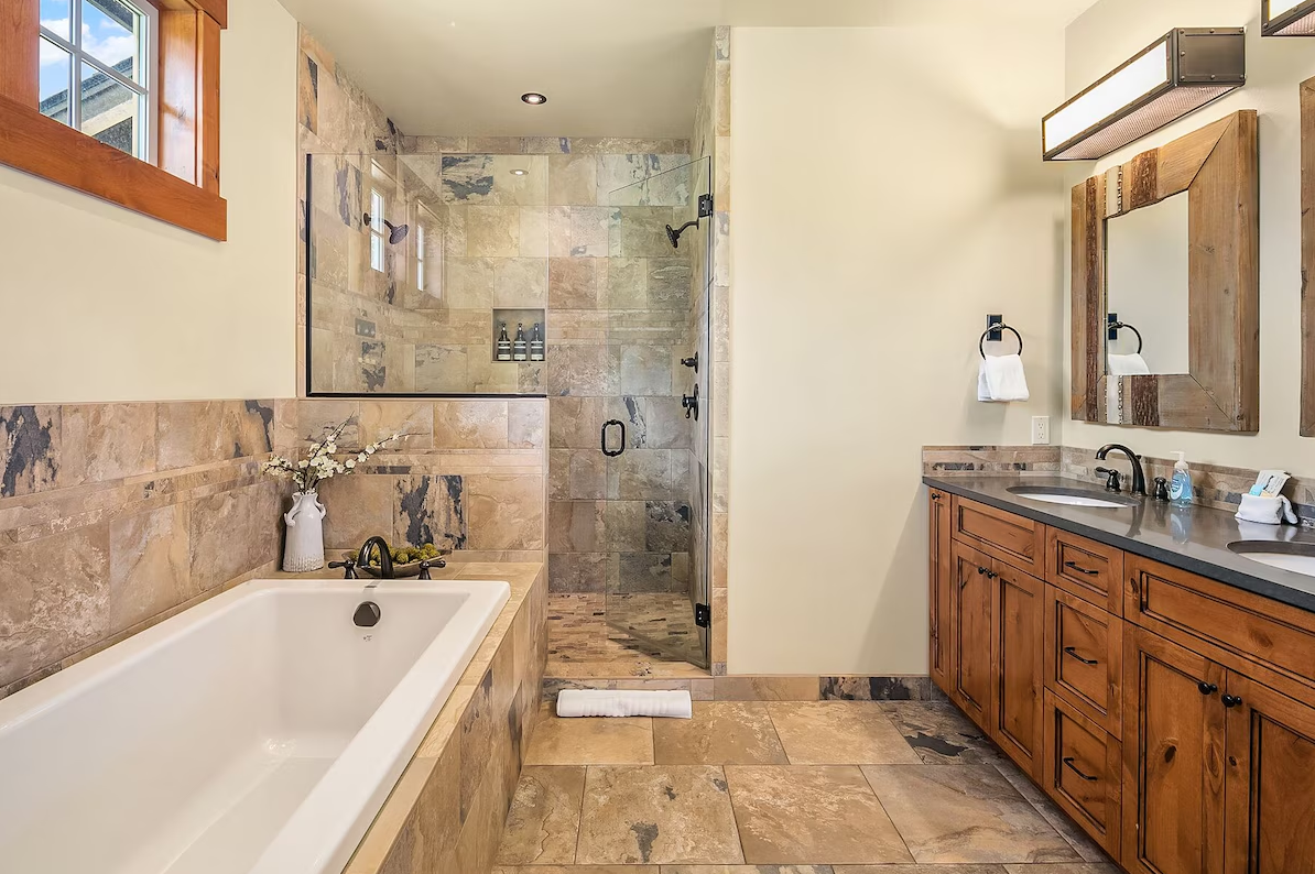 Bathroom with a bathtub, a glass-enclosed shower, wooden vanity with a double sink, two mirrors, and a window, featuring beige stone tiles.
