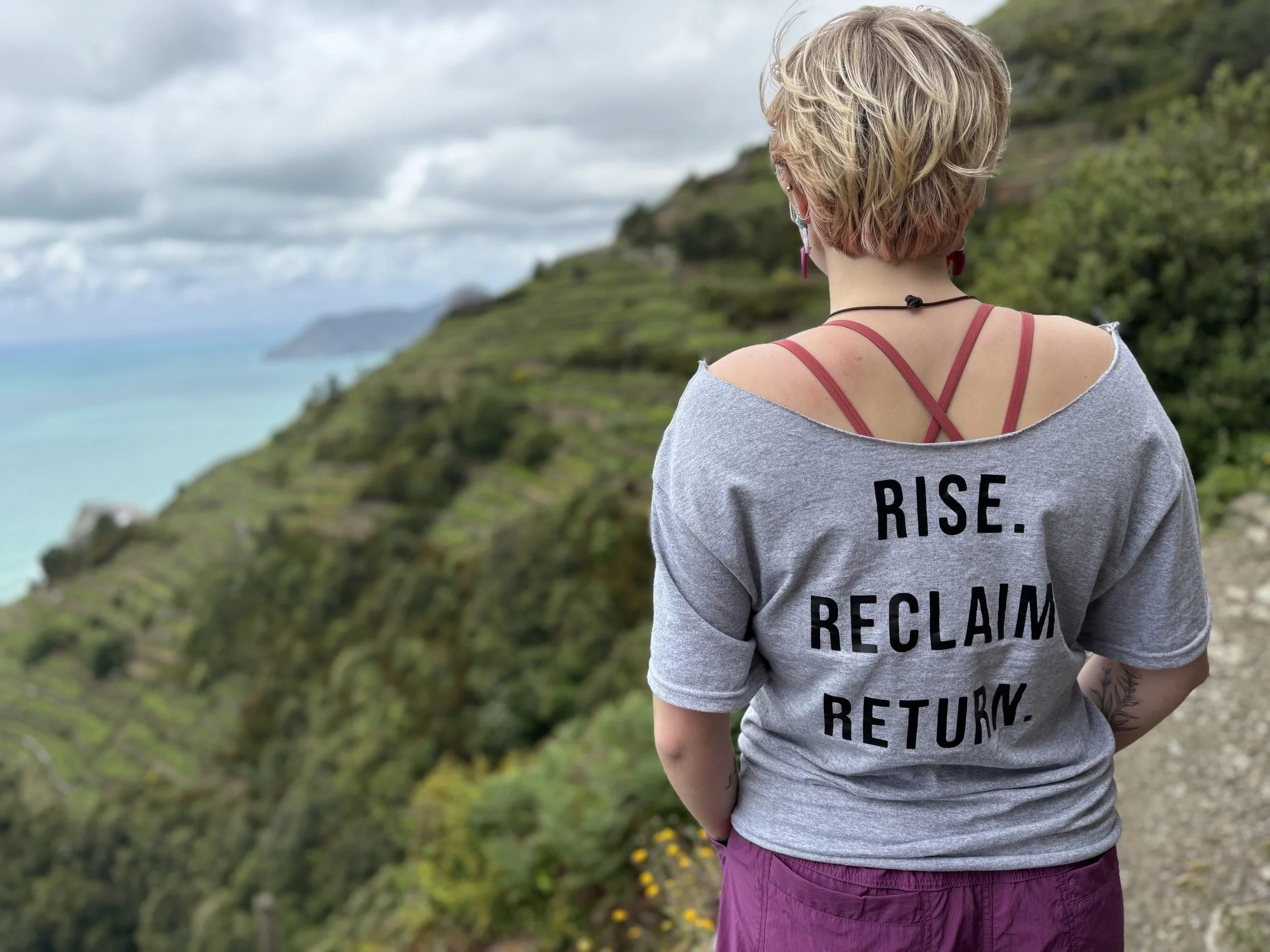 Back view of a woman with short blonde hair wearing a gray sweatshirt with the words 'Rise. Reclaim. Return.' on it, standing outdoors overlooking a lush green hillside and ocean in the distance under a cloudy sky.