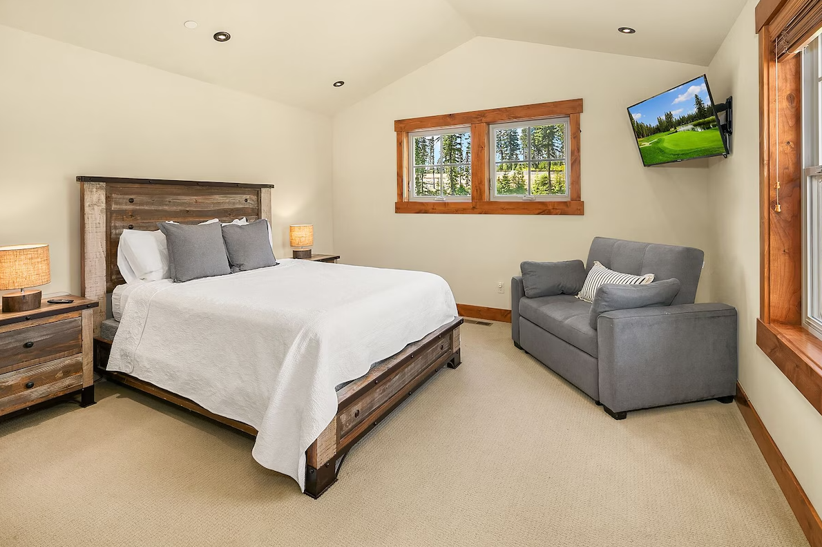 A bedroom with a wooden bed frame, white bedding, gray pillows, two wooden nightstands with lamps, two windows with wooden trim, a gray loveseat with pillows, and a wall-mounted TV showing a golf course.
