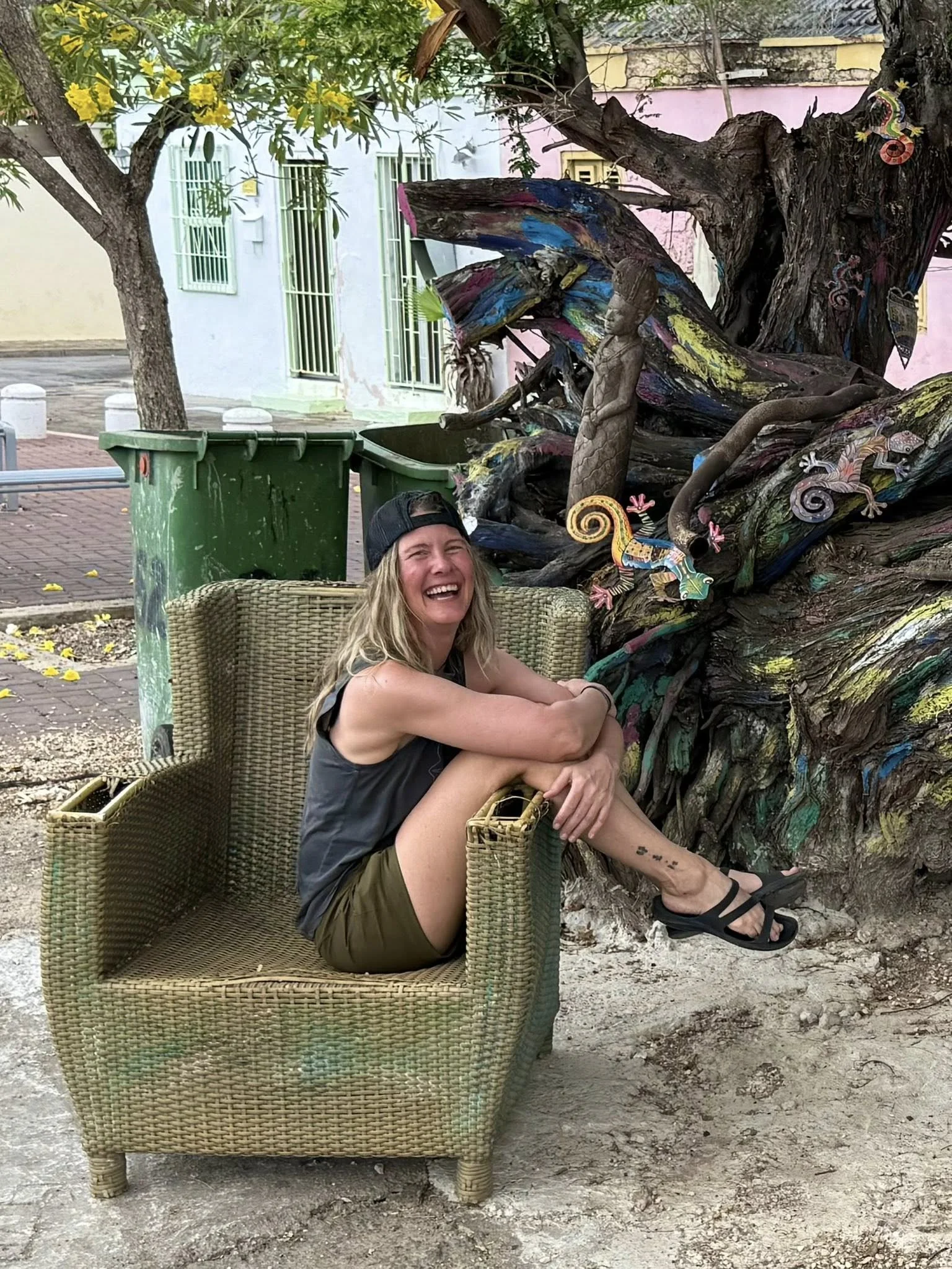 A person sitting in a wicker chair outdoors, smiling and laughing, with arms wrapped around knees, wearing a black cap, dark tank top, and shorts. In the background, there's a large, colorful, artistically painted tree with small painted lizards and 