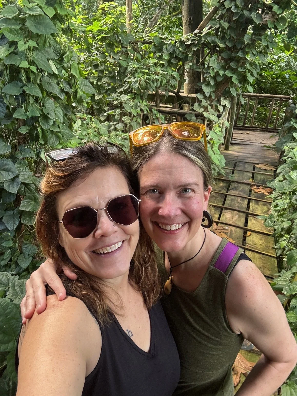 Two women smiling for a selfie outdoors in a lush green forest, with a wooden bridge in the background.