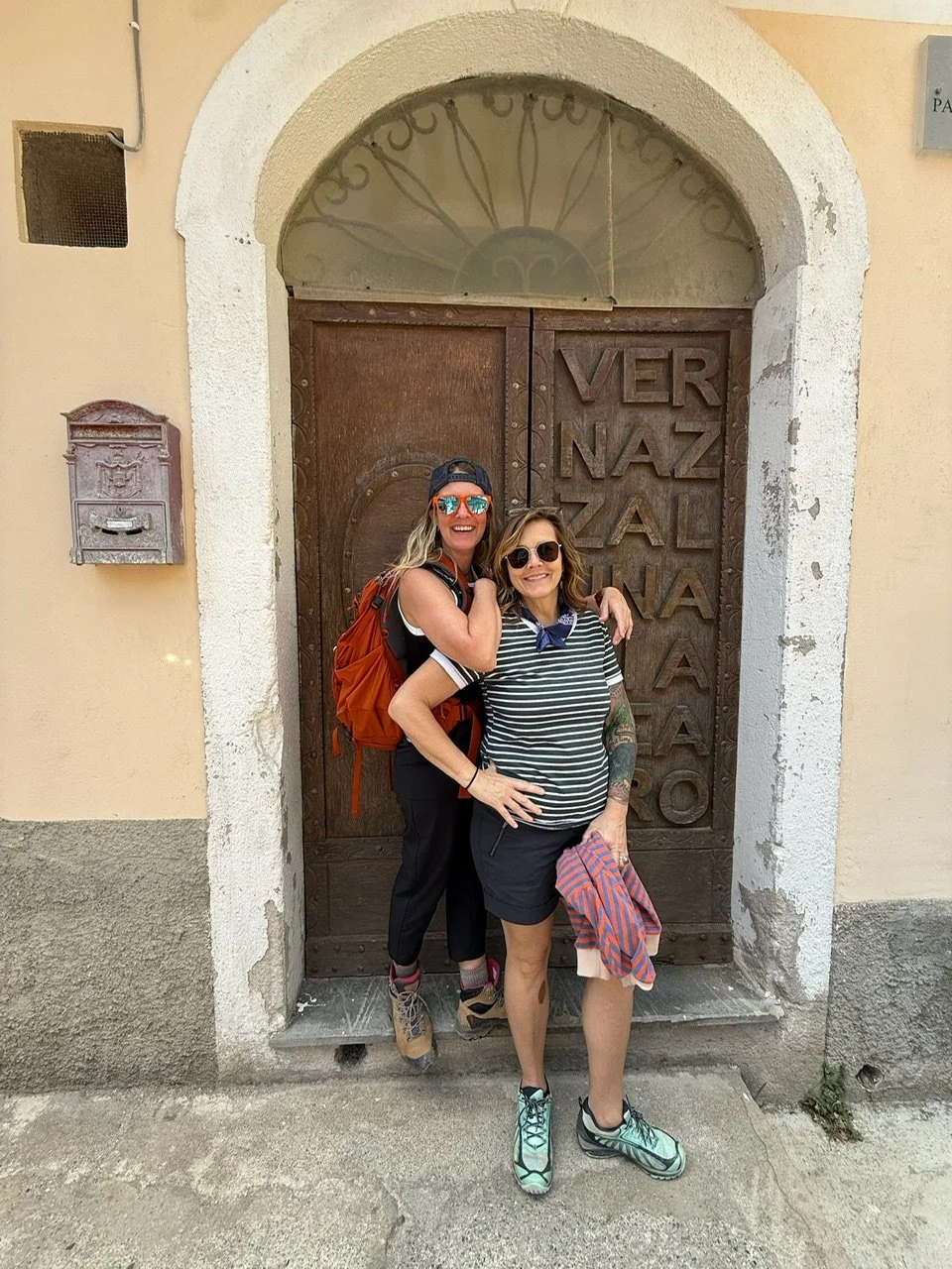 Two women smiling and posing together in front of a large wooden door with text, one with an orange backpack and hiking shoes, the other with tattoos and a striped shirt, both wearing sunglasses.