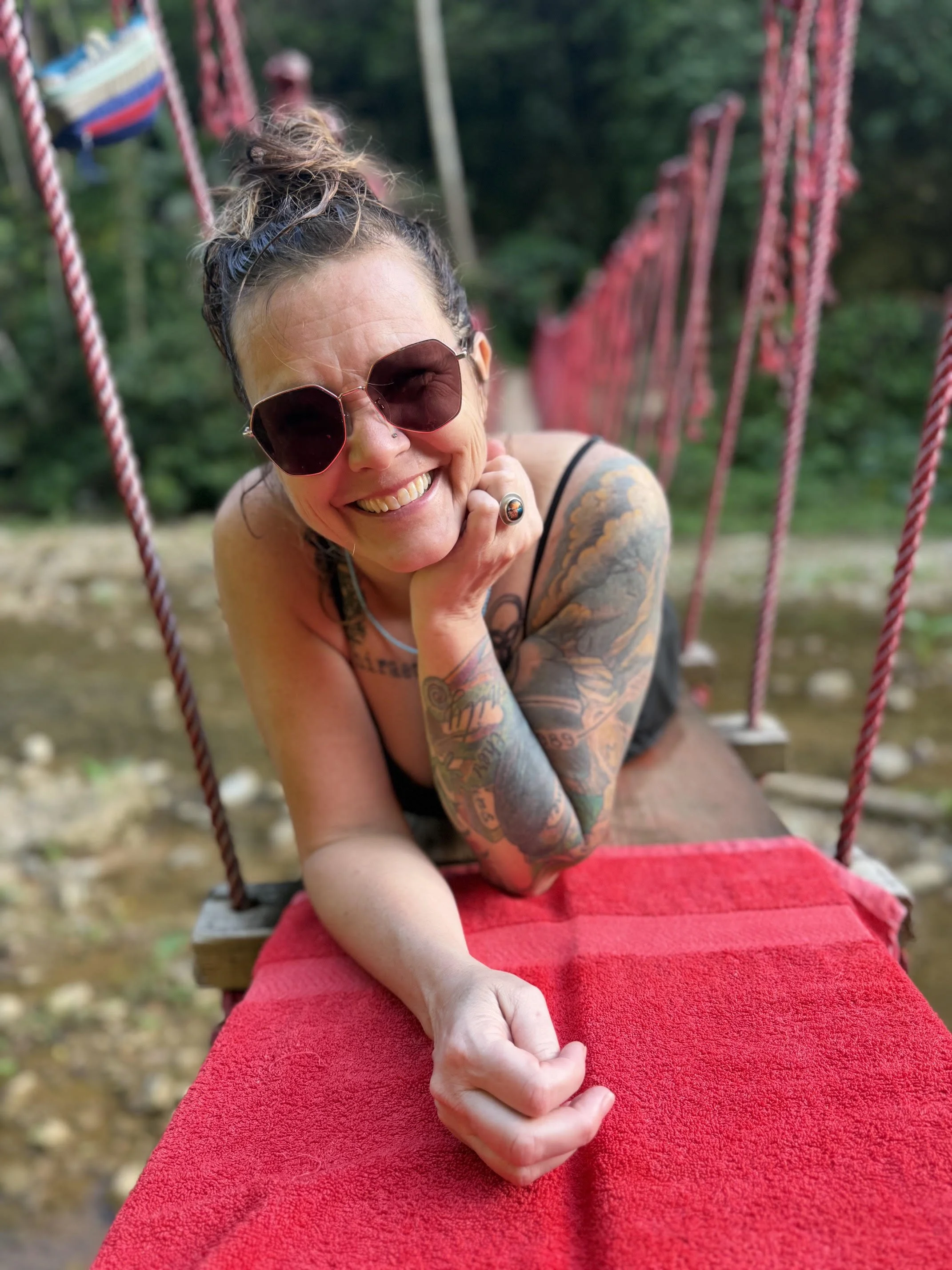 Smiling woman with tattoos, wearing sunglasses, sitting on a swing with a red towel, outdoors with blurred greenery in the background.