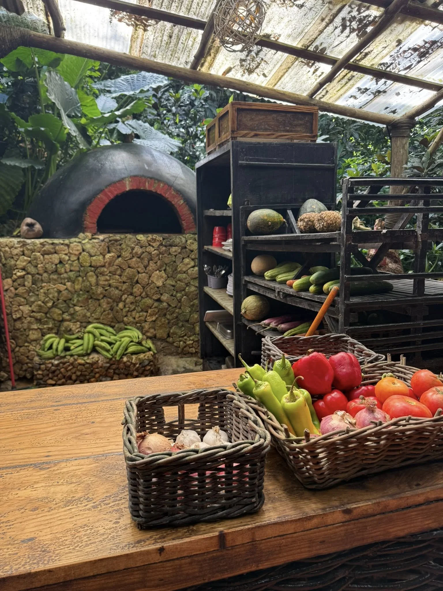 A wooden table with baskets of vegetables including green bell peppers, red bell peppers, tomatoes, and garlic. In the background, there is a rustic outdoor kitchen with a brick oven and a shelf with more vegetables and kitchen utensils. The setting 