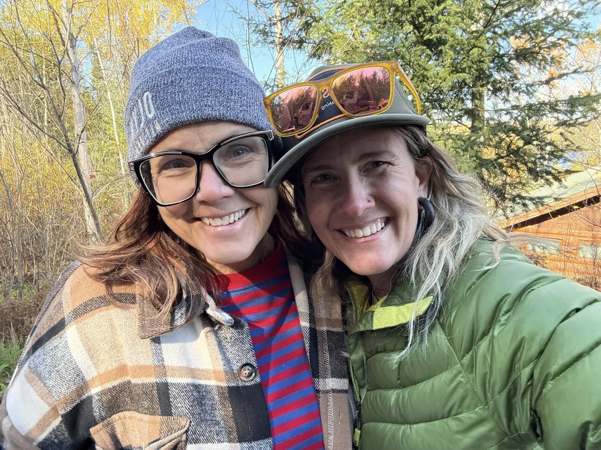 Two women smiling outdoors with trees and blue sky in the background. One wears a gray knit beanie, glasses, and a plaid jacket; the other wears a gray cap with sunglasses on top and a green jacket.