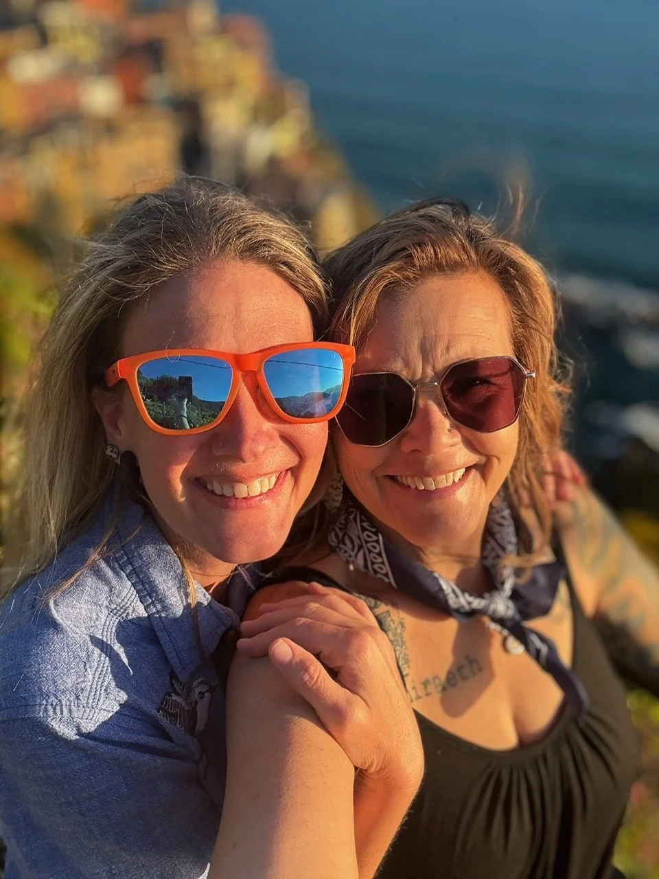 Two women taking a selfie with a colorful coastline and ocean in the background during sunset.