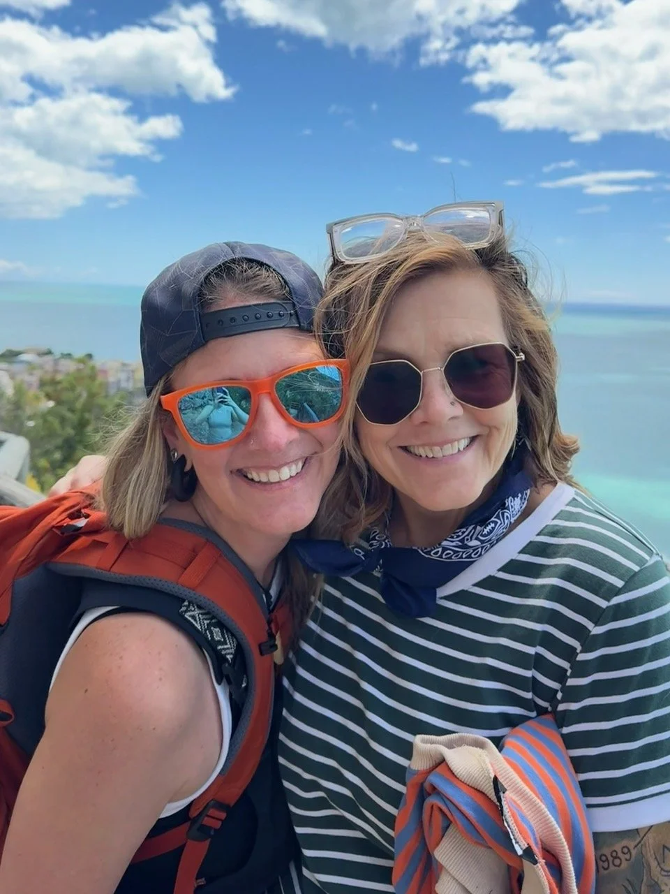 Two women smiling outdoors with ocean and sky in the background, wearing sunglasses and casual clothing. Chris Meland. Julie "Jitterbug" Pearce