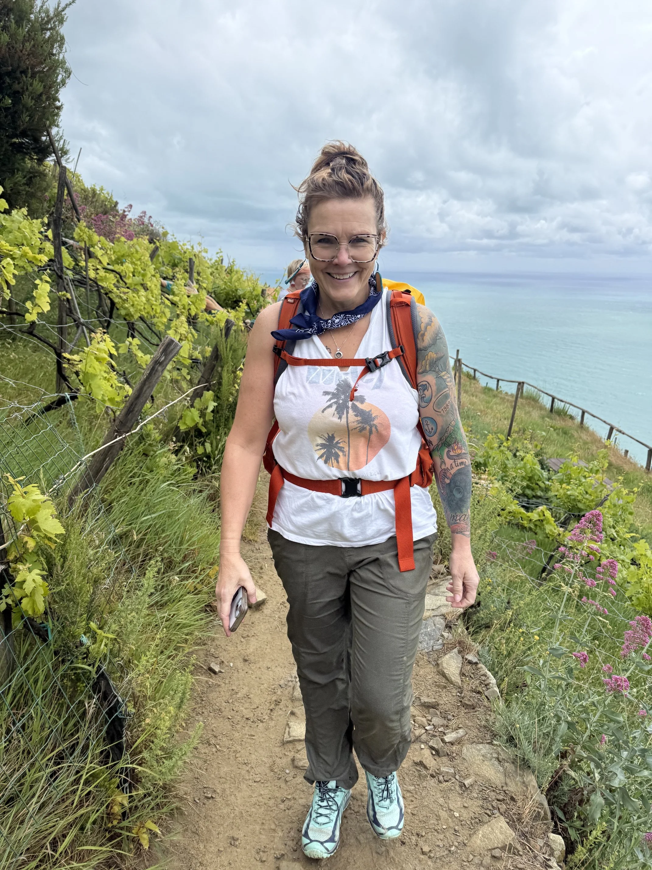 A smiling woman with glasses and tattoos on her arms, wearing a white tank top with a palm tree sunset design, gray pants, turquoise hiking shoes, an orange backpack, and a blue bandana, walking on a dirt trail along a coastline with green plants and