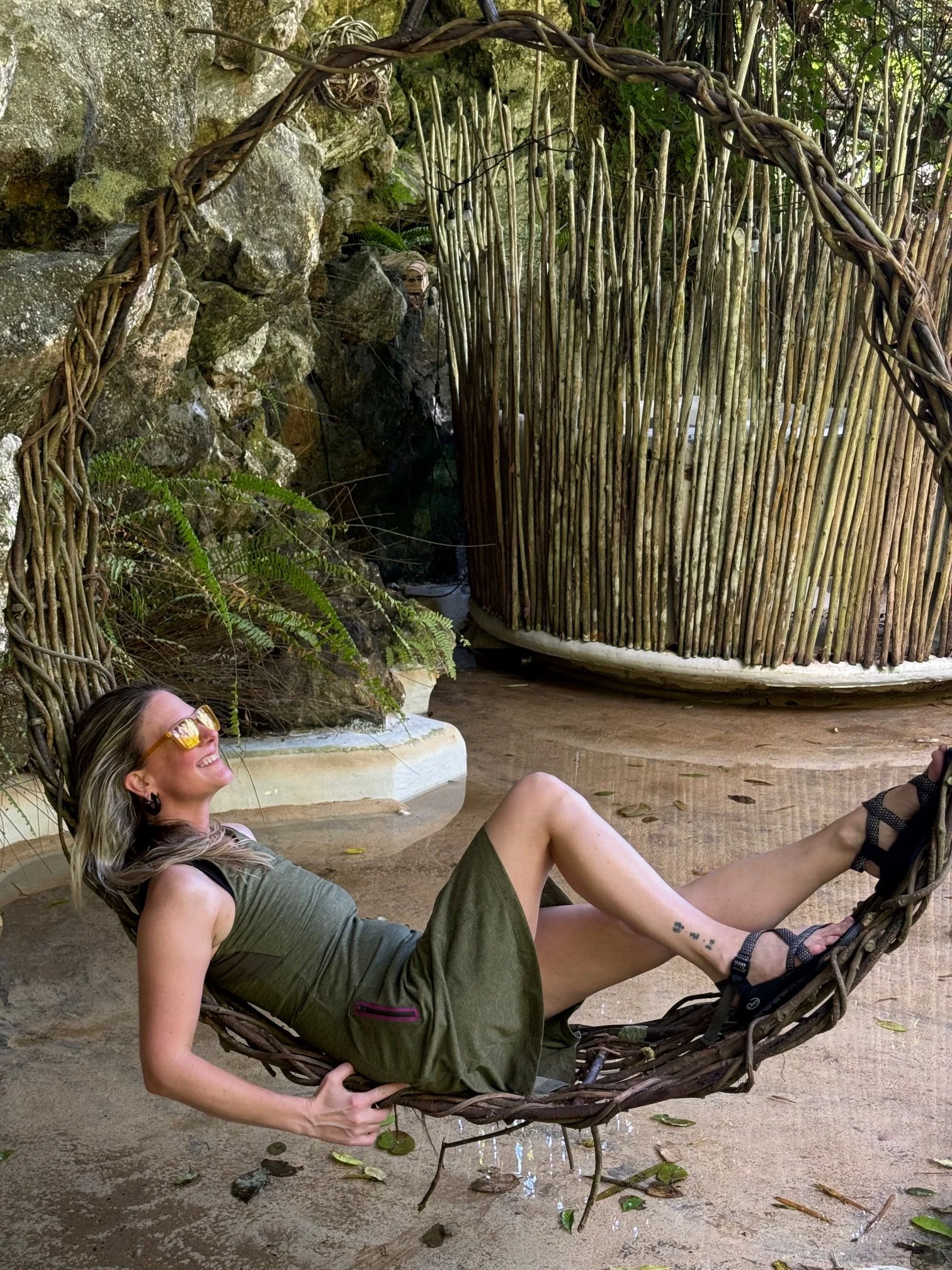 A woman with long blonde hair, wearing sunglasses, a gray sleeveless top, and green shorts, is reclining and smiling in a hanging wicker basket in a natural outdoor setting with rocks, greenery, and a sandy ground.