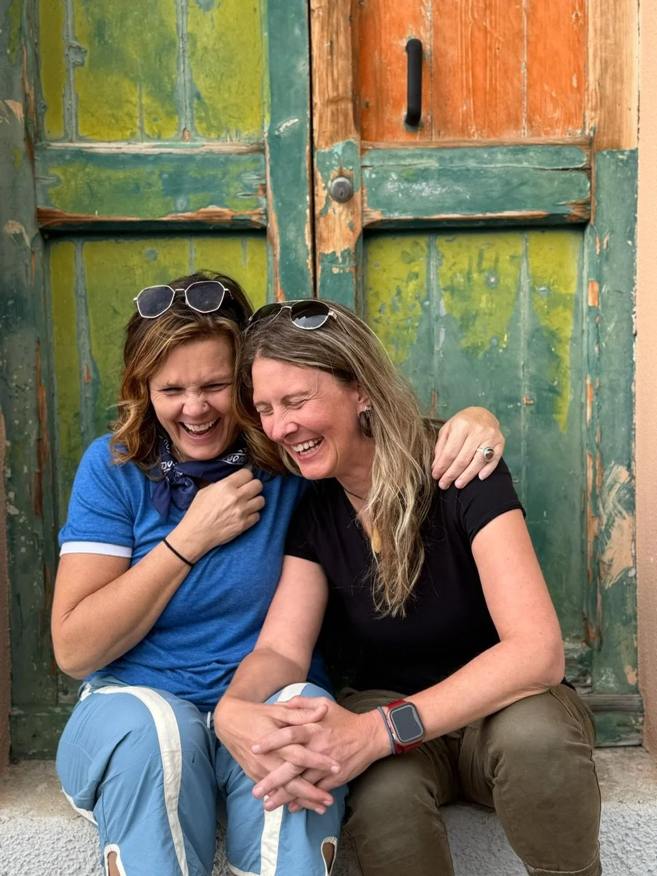 Two women are sitting close together outdoors, smiling and laughing. They are embracing each other with their heads touching, and the background is a weathered green and orange wooden door. Chris Meland. Julie "Jitterbug" Pearce