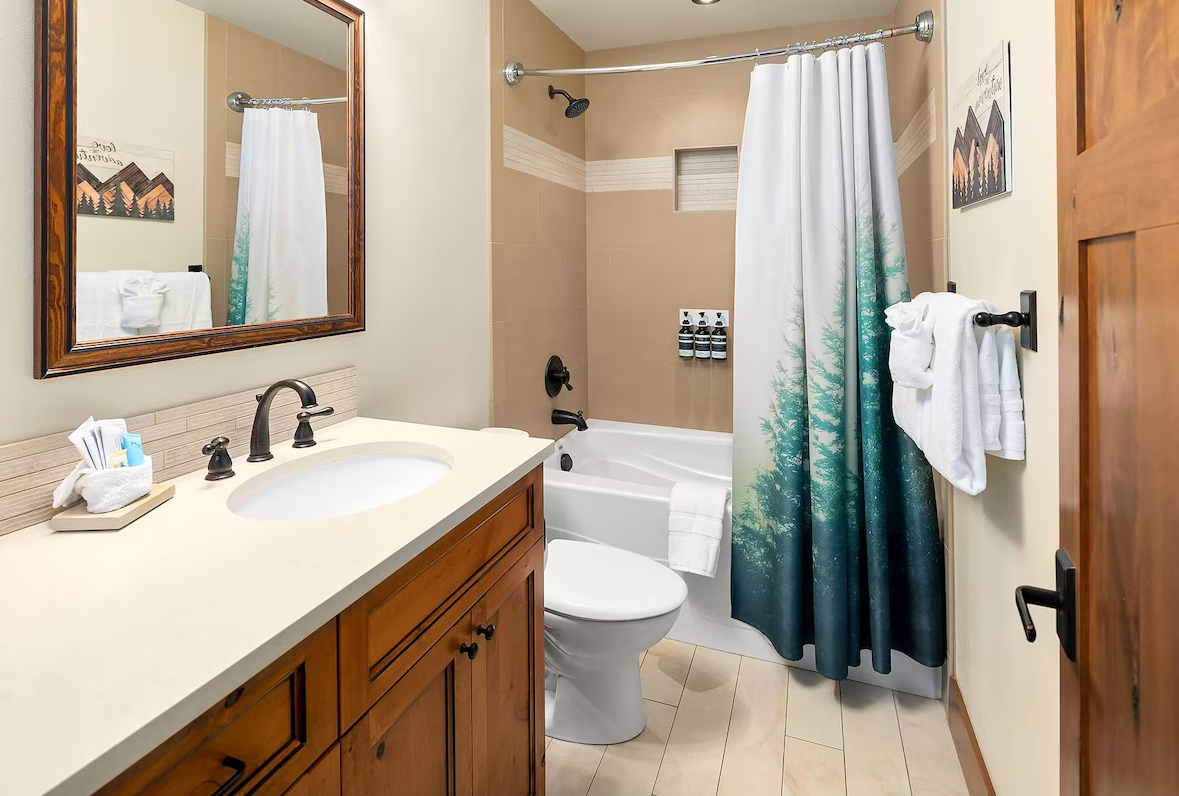 A bathroom with a wooden vanity, mirror, and sink, next to a bathtub with a shower curtain that depicts a forest scene. There are towels hanging on a black towel rack and bathroom amenities on a wall-mounted shelf in the shower area.