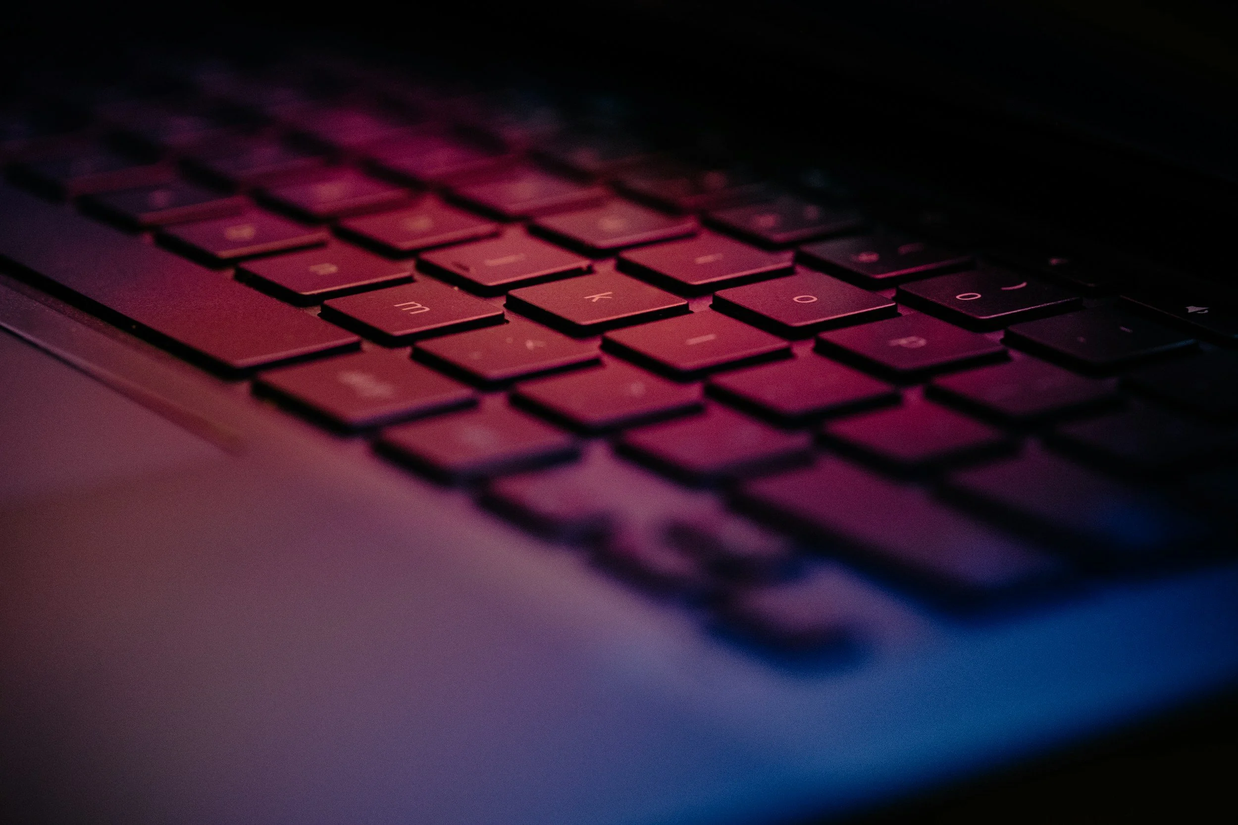 A close up angled shot of a computer keyboard with pink and blue lighting on it