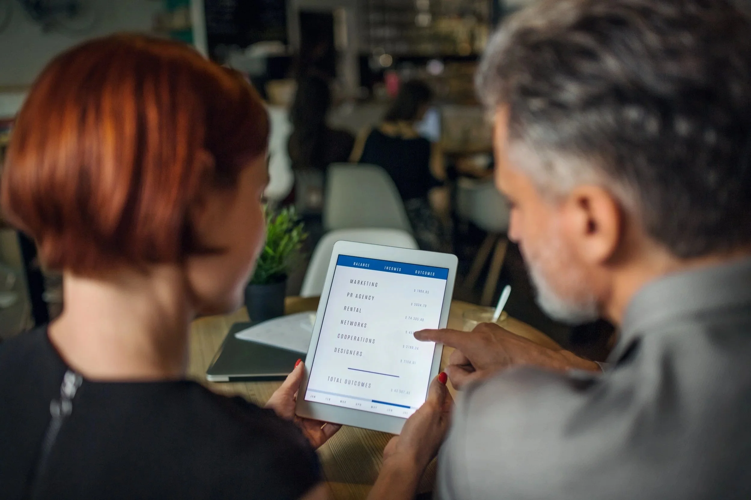 Two people in a cafe looking at a tablet with financial data on the screen, with other patrons blurred in the background.