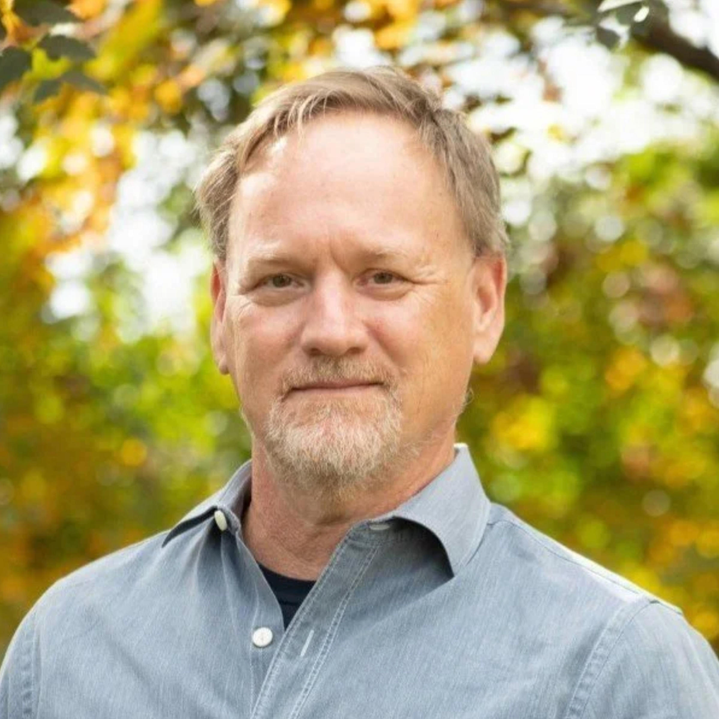 A middle-aged man with light hair, beard, and mustache posing outdoors with autumn leaves in the background.