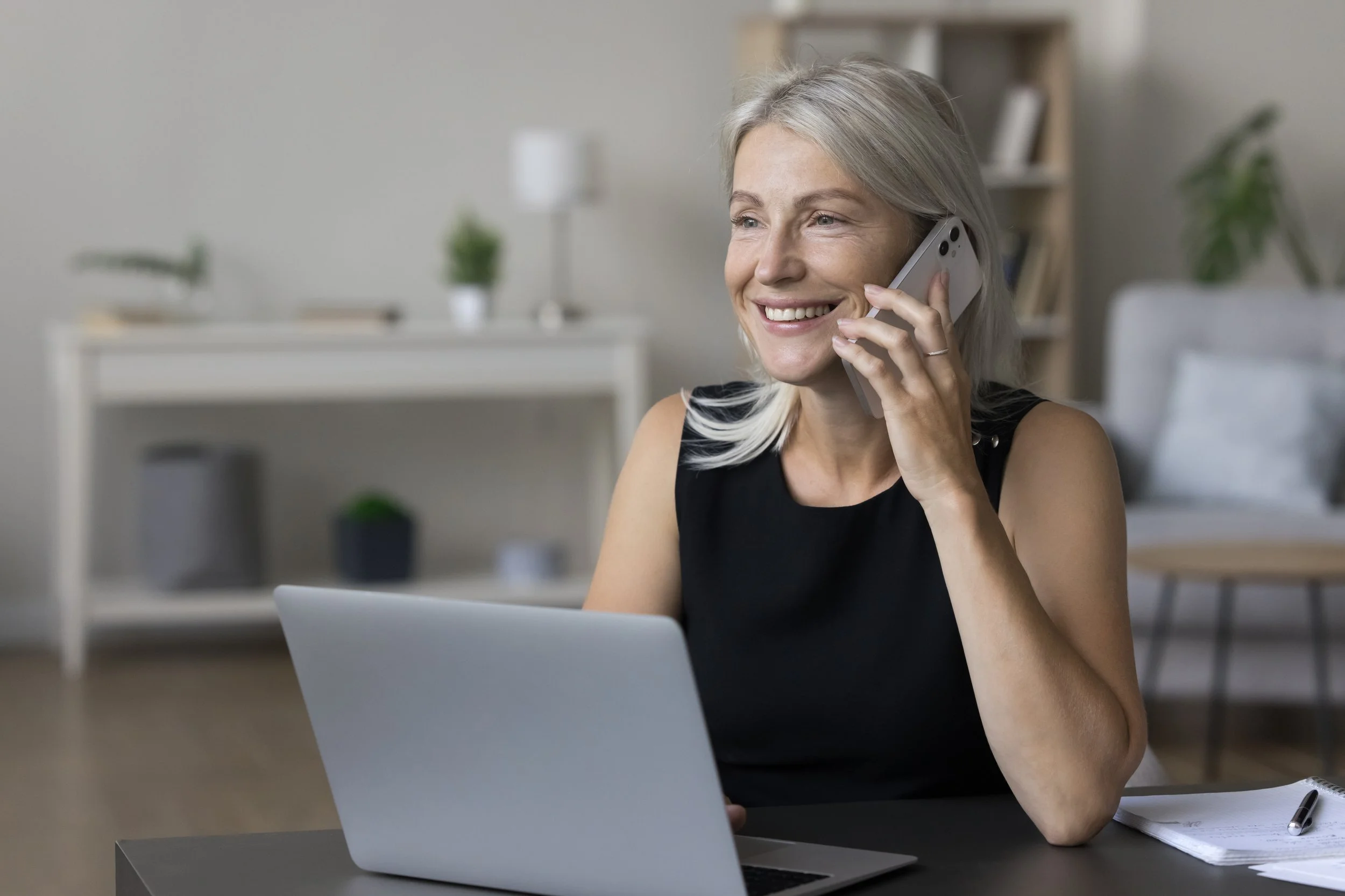 A smiling woman with gray hair, wearing a black sleeveless top, talking on a smartphone while working on a laptop at a desk in a bright living room.