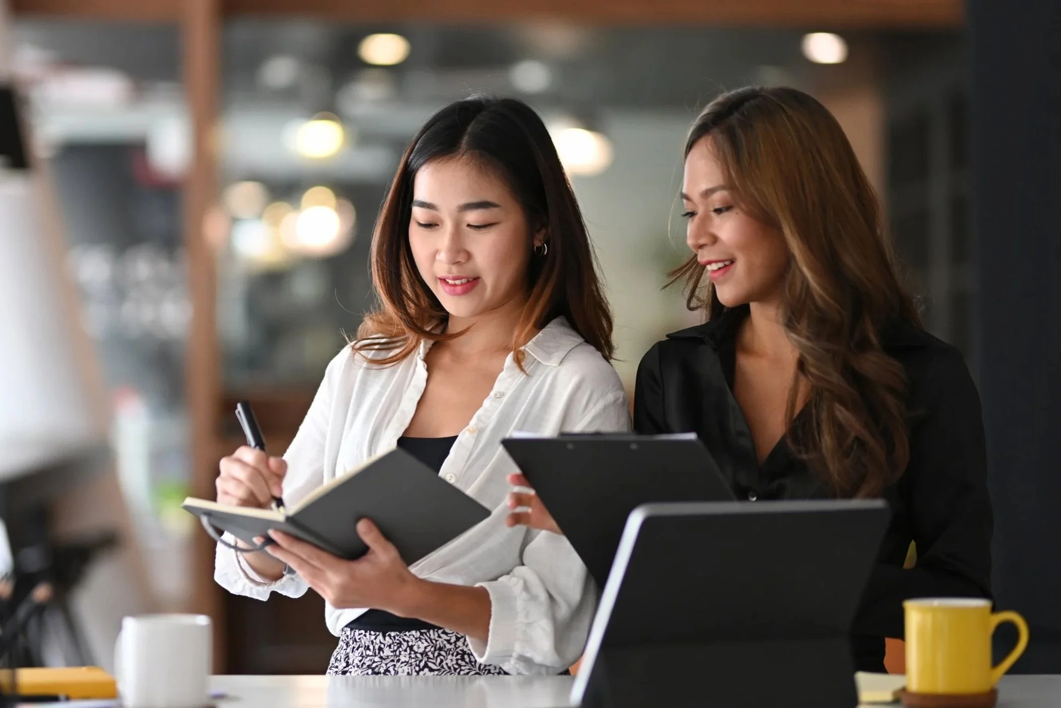 Two women standing in a cafe, looking at notebooks and smiling with a laptop and coffee mugs on the table.