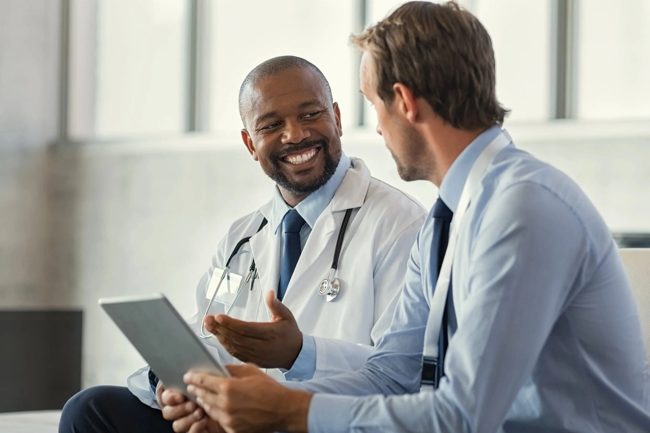 Two male doctors in white coats sitting and talking, one holding a tablet, the other smiling, in a hospital or clinic setting.