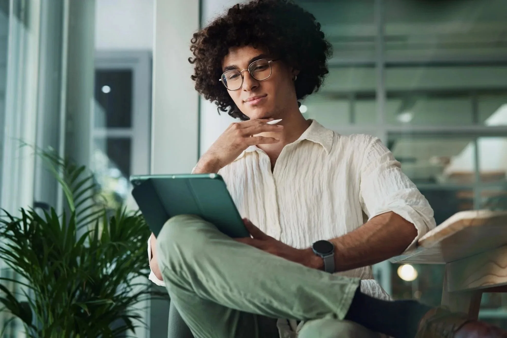 Young man with curly hair and glasses sitting by a window, reading a tablet, with a plant nearby.