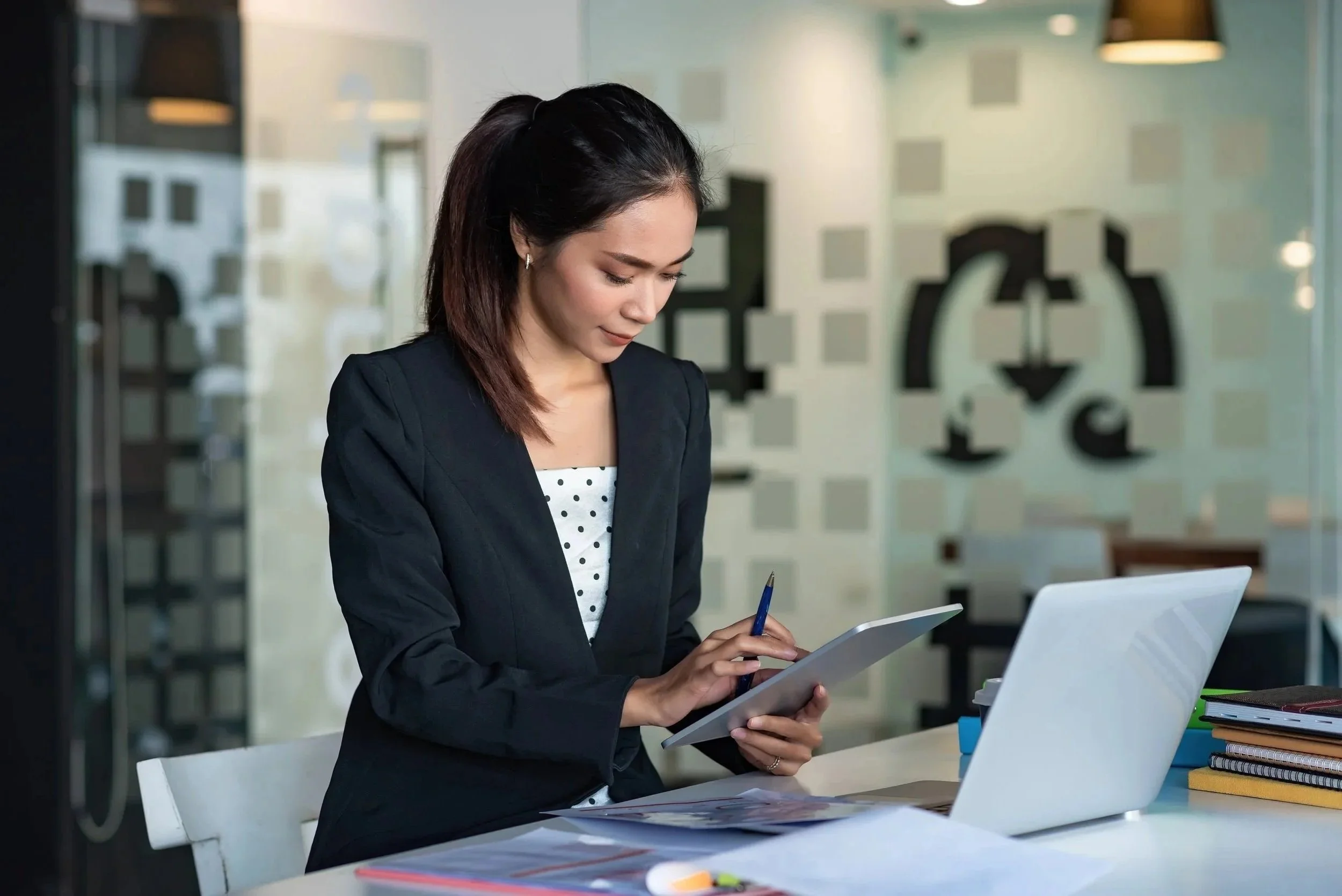 A woman with dark hair in a professional black blazer and white polka dot top is standing in an office, looking down and working on a digital tablet with a stylus. On the desk in front of her are a white laptop, colorful folders, and a notebook.