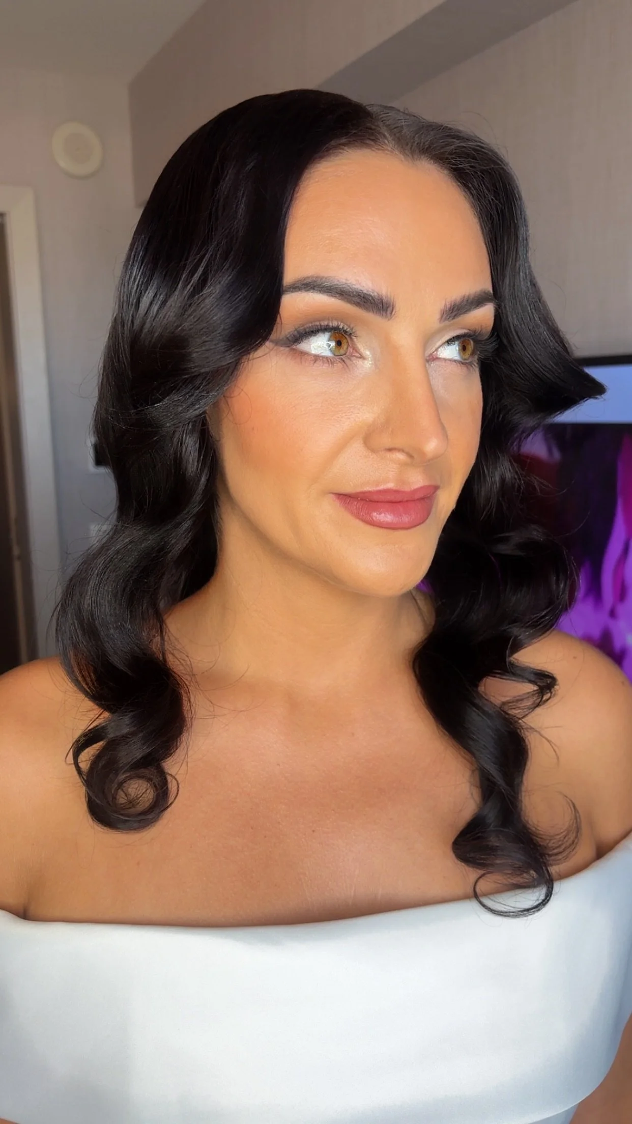 A woman with dark wavy hair, light makeup, and a white off-shoulder top, looking slightly to the right.