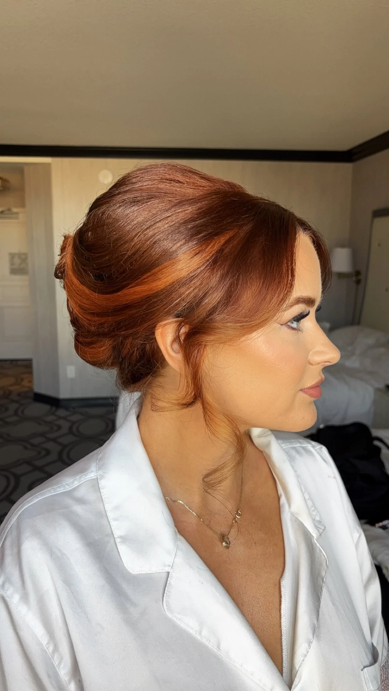 A woman with styled auburn hair in an elegant updo, wearing a white shirt and a delicate necklace, in a hotel room.