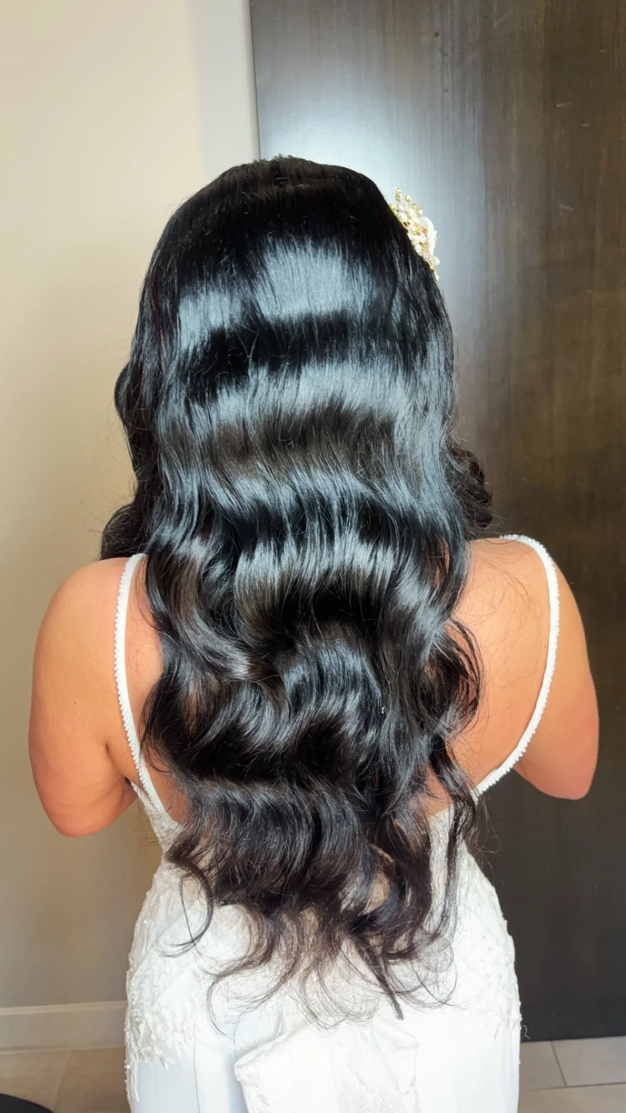 Back view of woman with long, shiny, wavy black hair wearing a white dress with thin straps and decorative elements.