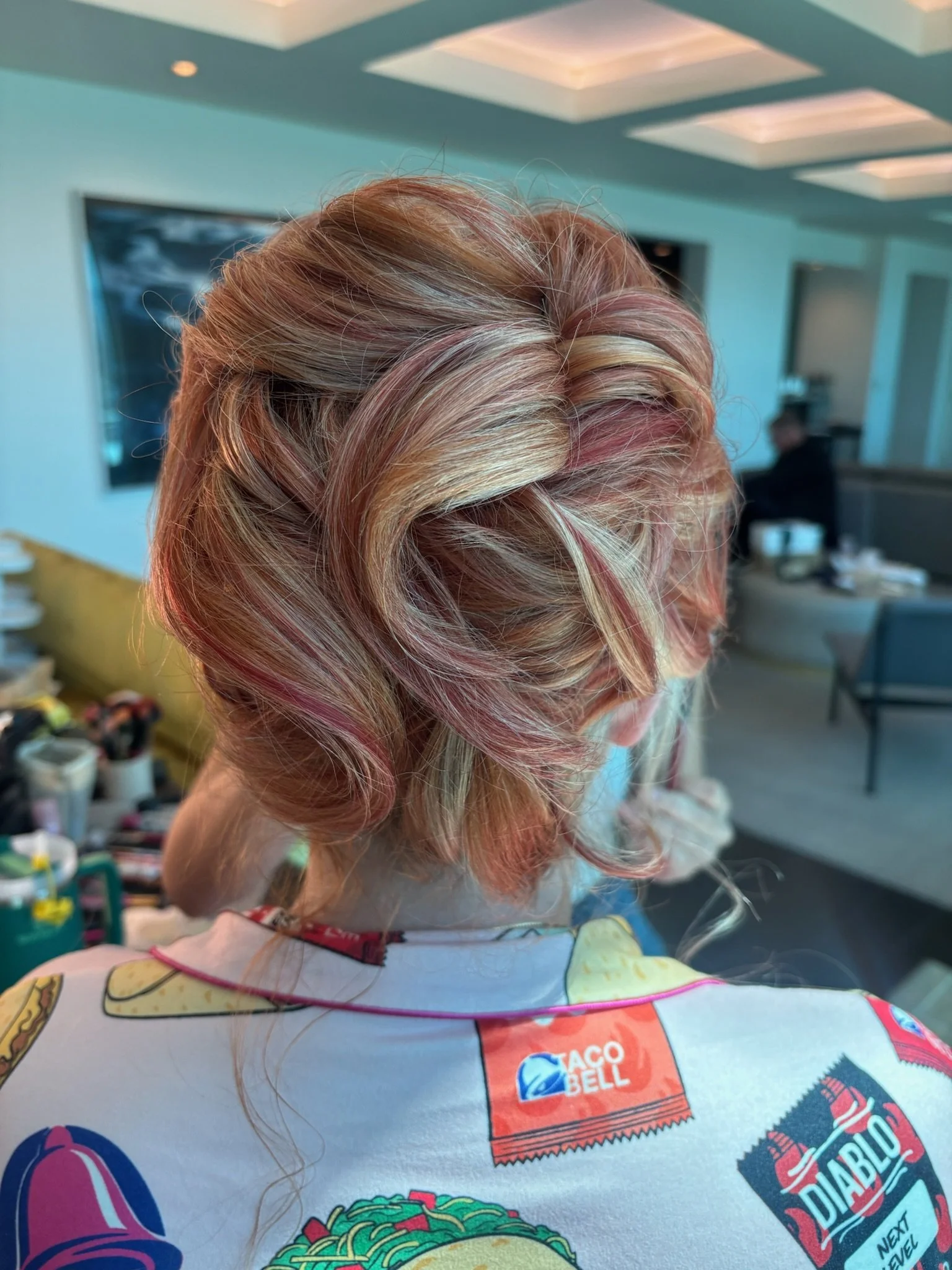 A woman with multicolored, styled hair in shades of blonde, pink, and red, viewed from behind in a room with modern decor.