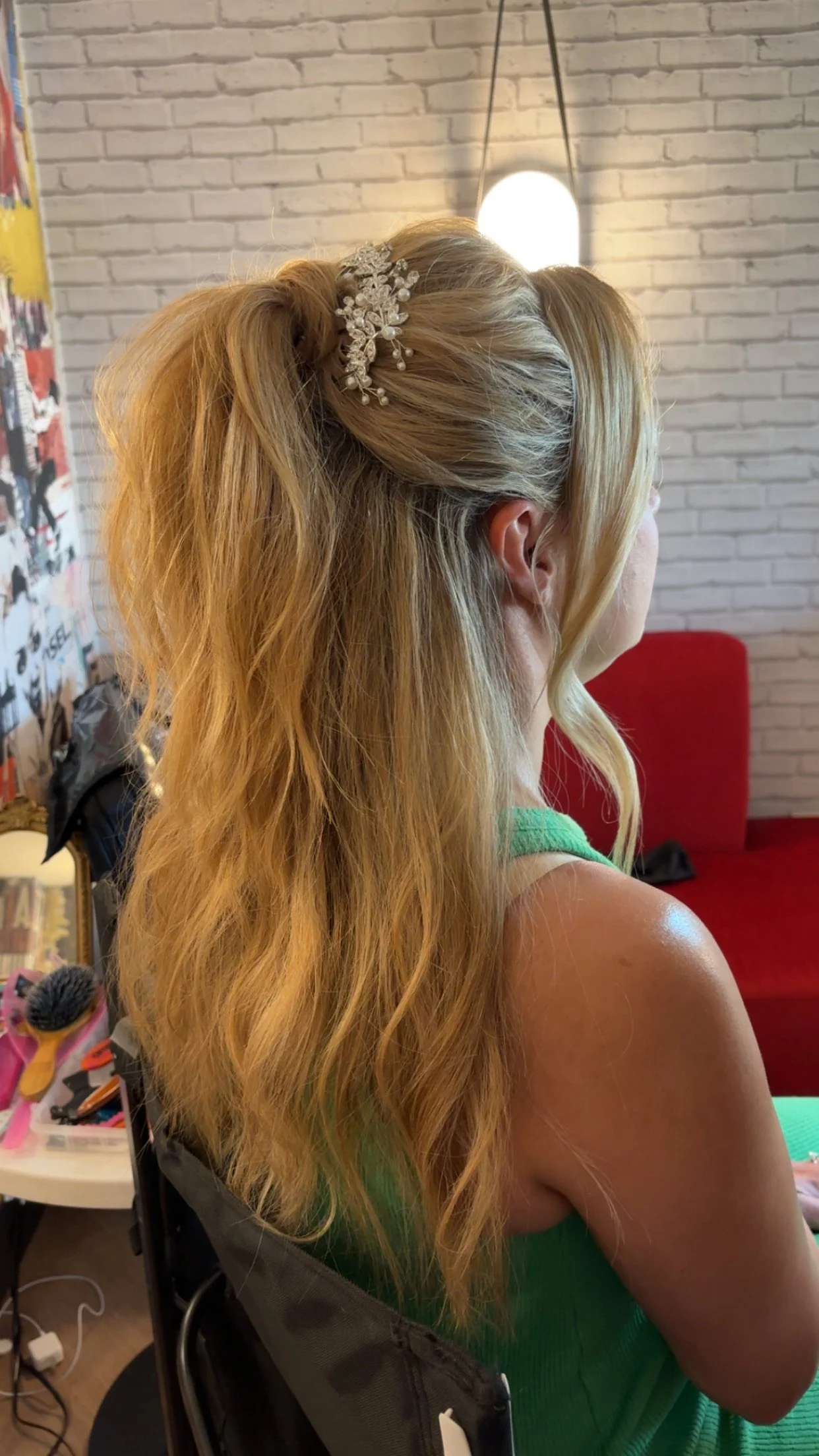 Side view of a woman with long, wavy blonde hair styled in a half-updo, secured with a decorative hair clip, sitting in a room with a white brick wall and a red couch.