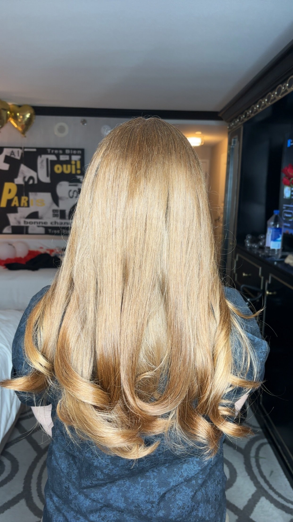 Back view of a woman with long, blonde, styled hair with soft curls, standing in a room with a black and gold decor, a wall with black and white posters, and a TV.
