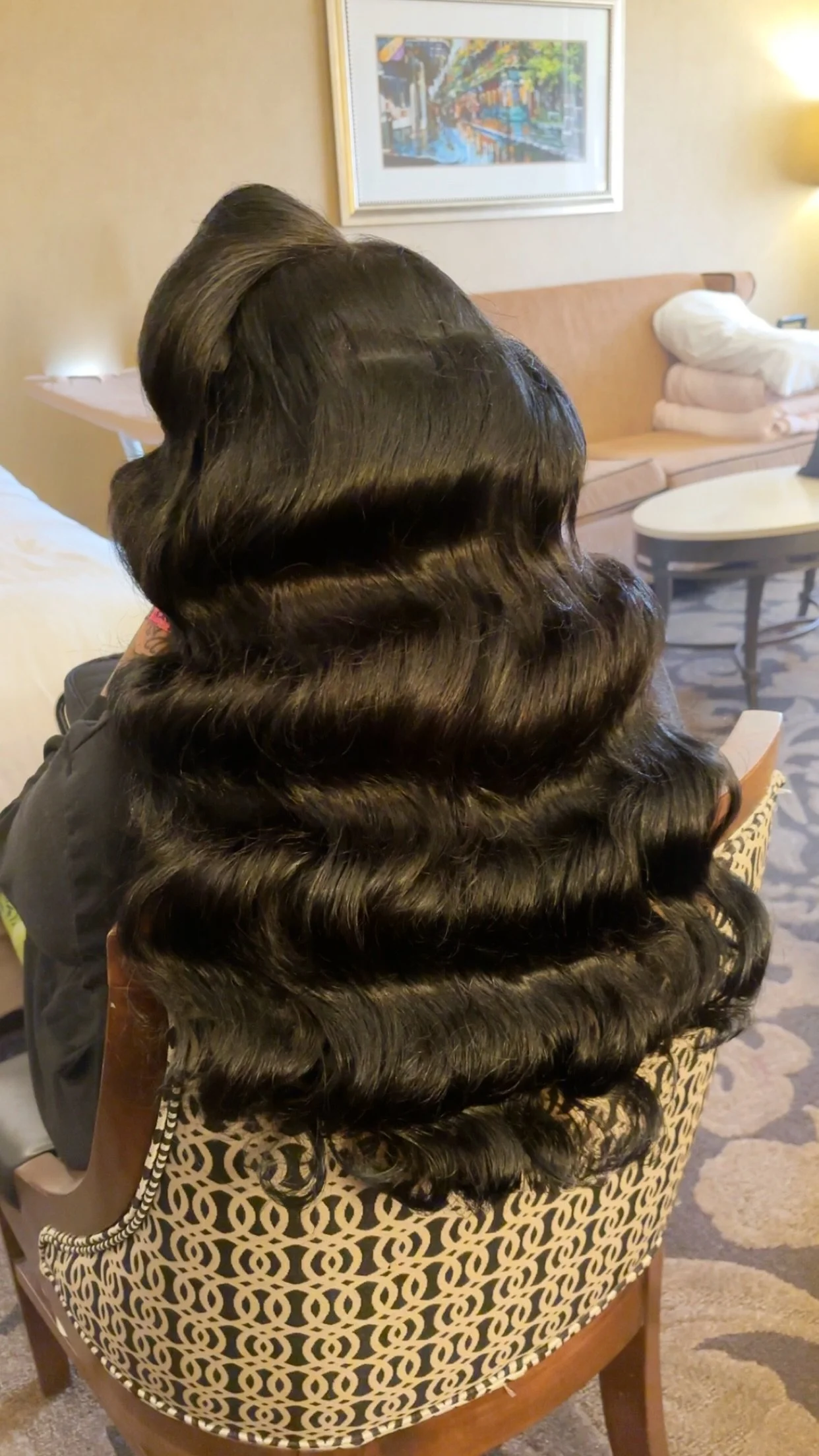 Close-up of a woman with long, shiny, dark, styled hair with large, defined curls sitting on a patterned chair in a hotel room.