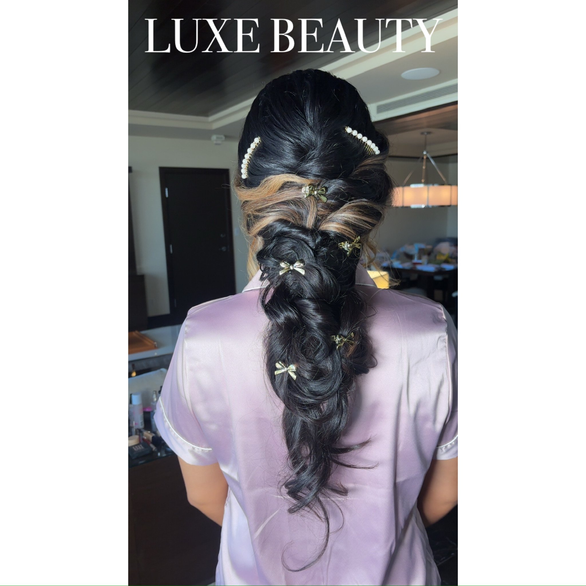 Back of a woman's head with long dark hair styled in a loose braid decorated with small gold bows and pearls, wearing a shiny pink satin robe, with the words 'LUXE BEAUTY' at the top.