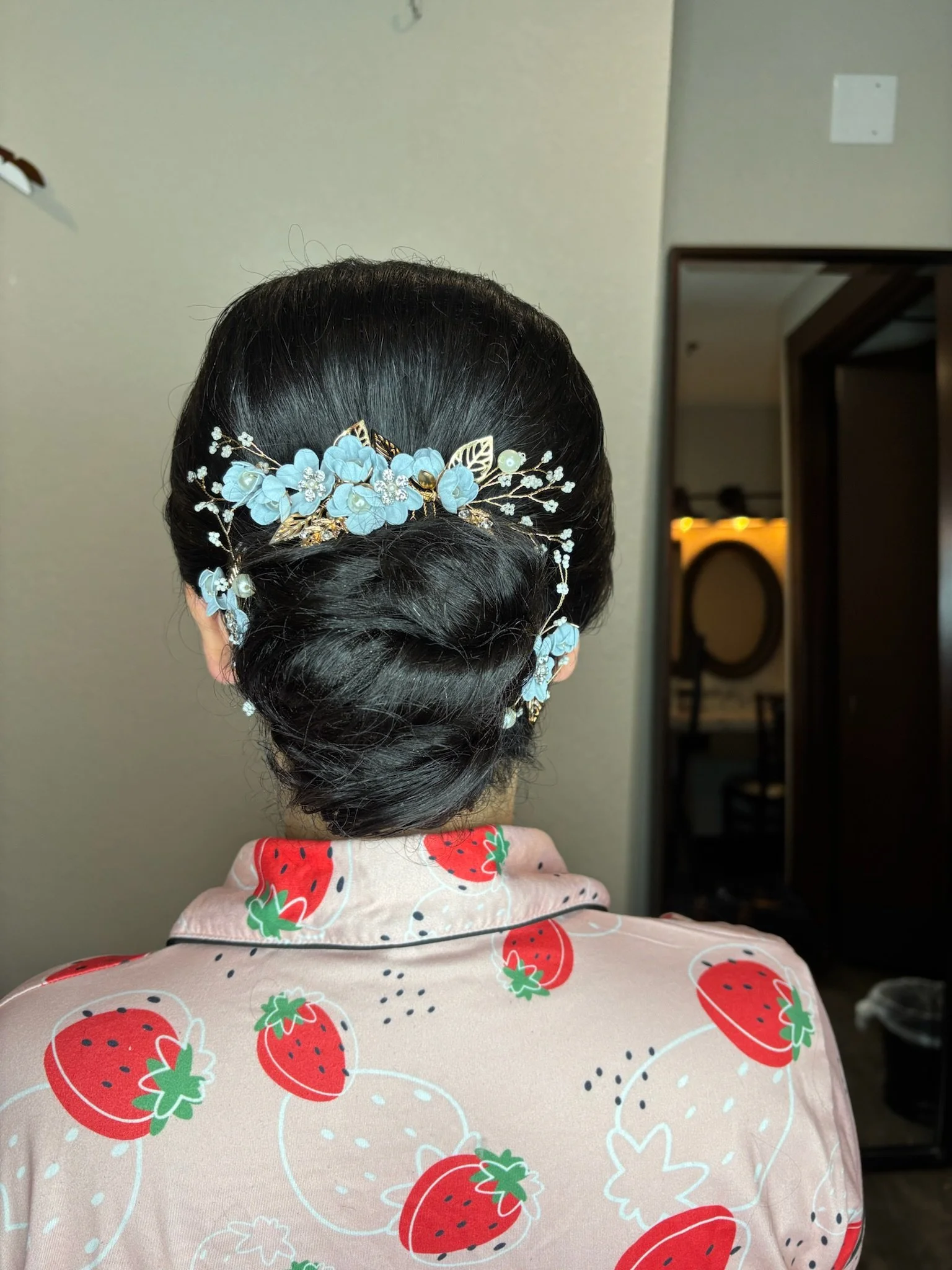 Back view of a woman with dark hair styled in an elegant updo, decorated with a light blue floral hairpiece with pearls and gold accents, wearing a pink pajama top with red strawberry patterns.