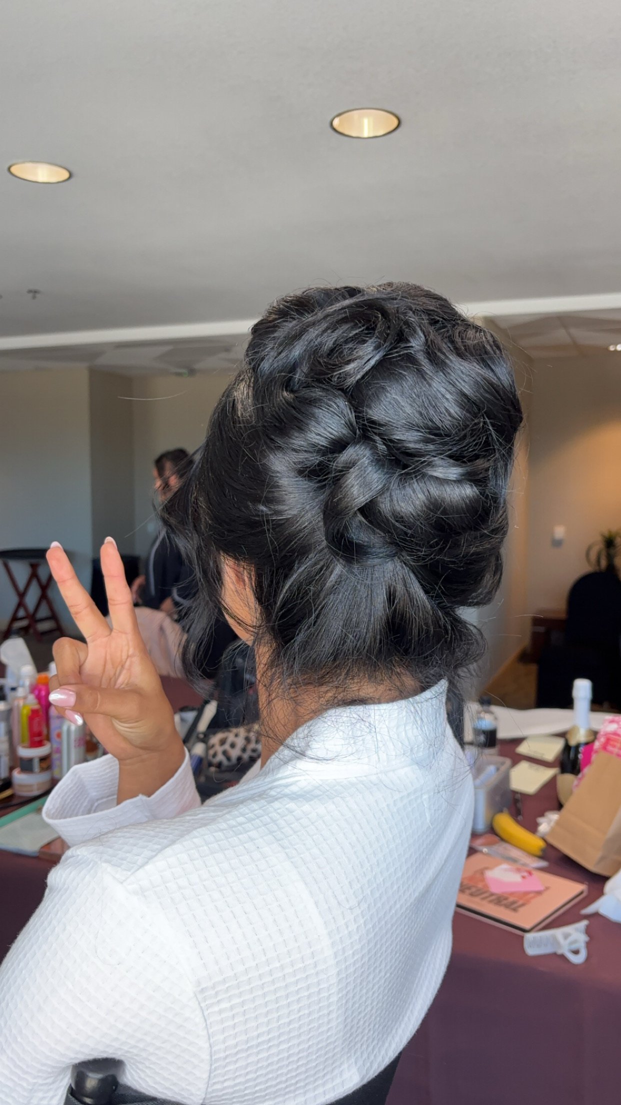 Back view of a woman with styled black hair in an updo, making a peace sign with her right hand, wearing a white textured blouse, in a room with various hairstyling tools and products on a table.