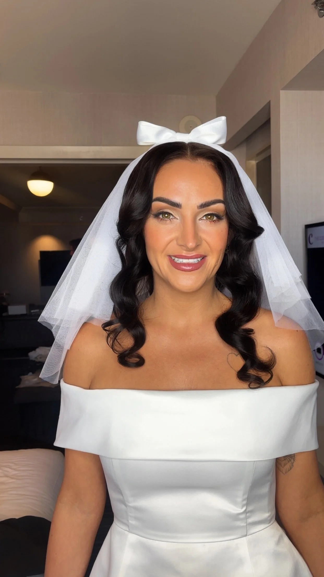 A woman with dark, wavy hair wearing a white off-the-shoulder dress and a bridal veil with a large white bow on top, inside a room.