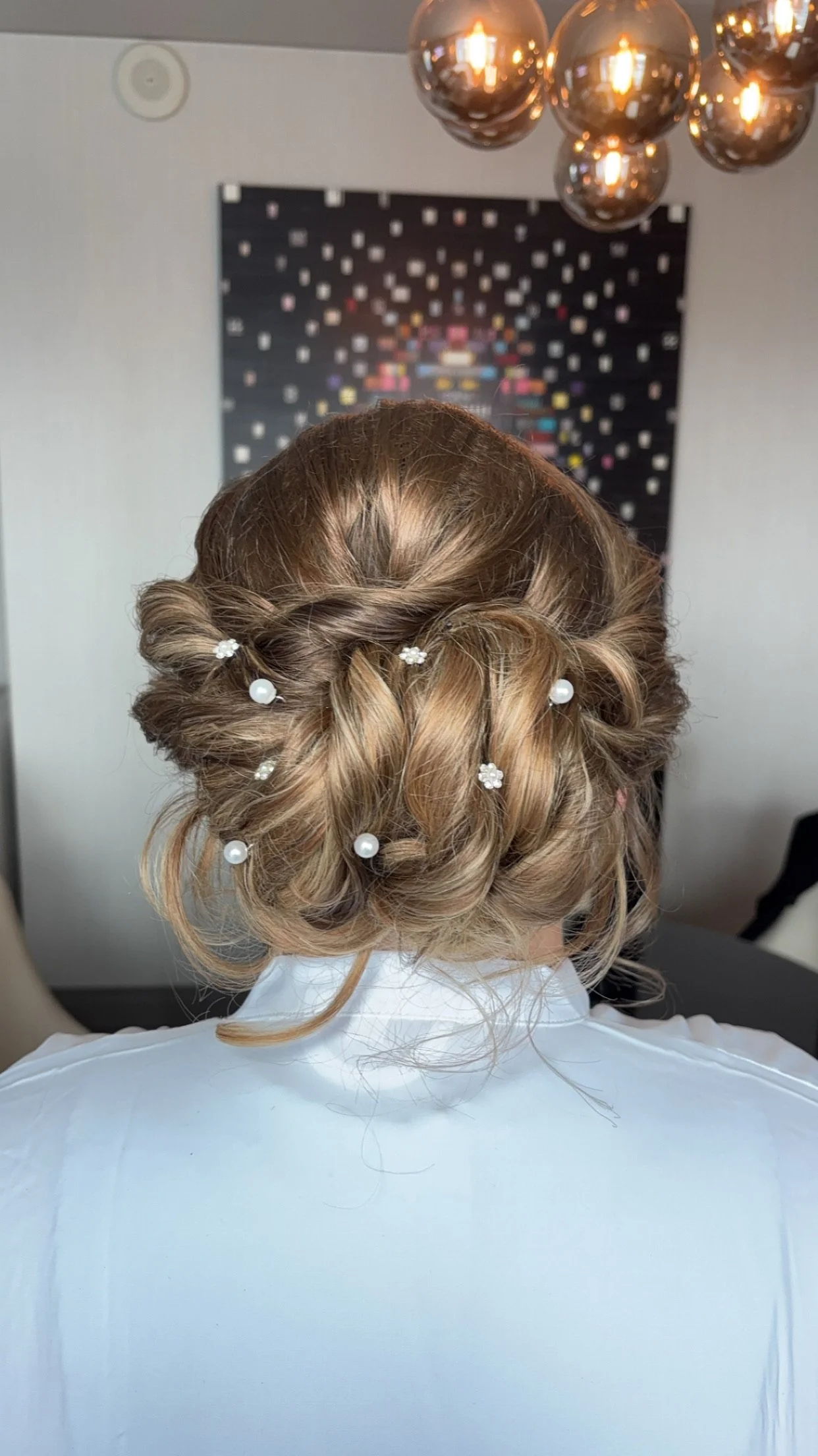 Back of a woman's head with a braided updo hairstyle decorated with pearl and flower hairpins, wearing a white shirt, in a room with a modern chandelier and abstract art on the wall.