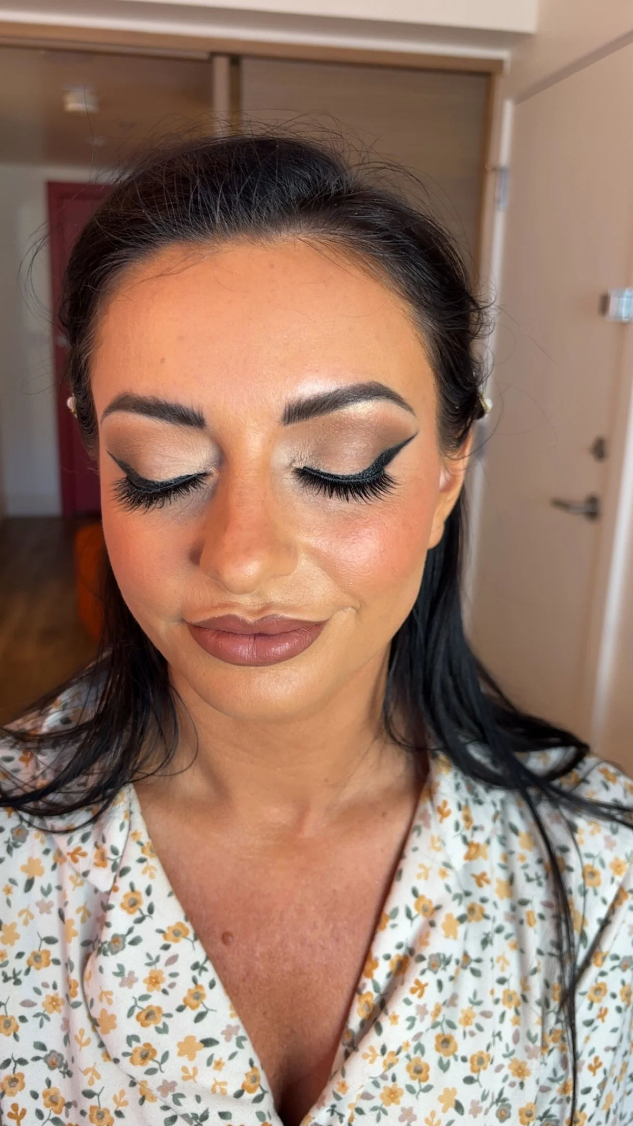 A woman with dark hair, closed eyes, and makeup with winged eyeliner, eyeshadow, blush, and lipstick, wearing a floral top, indoors with wooden and beige walls.